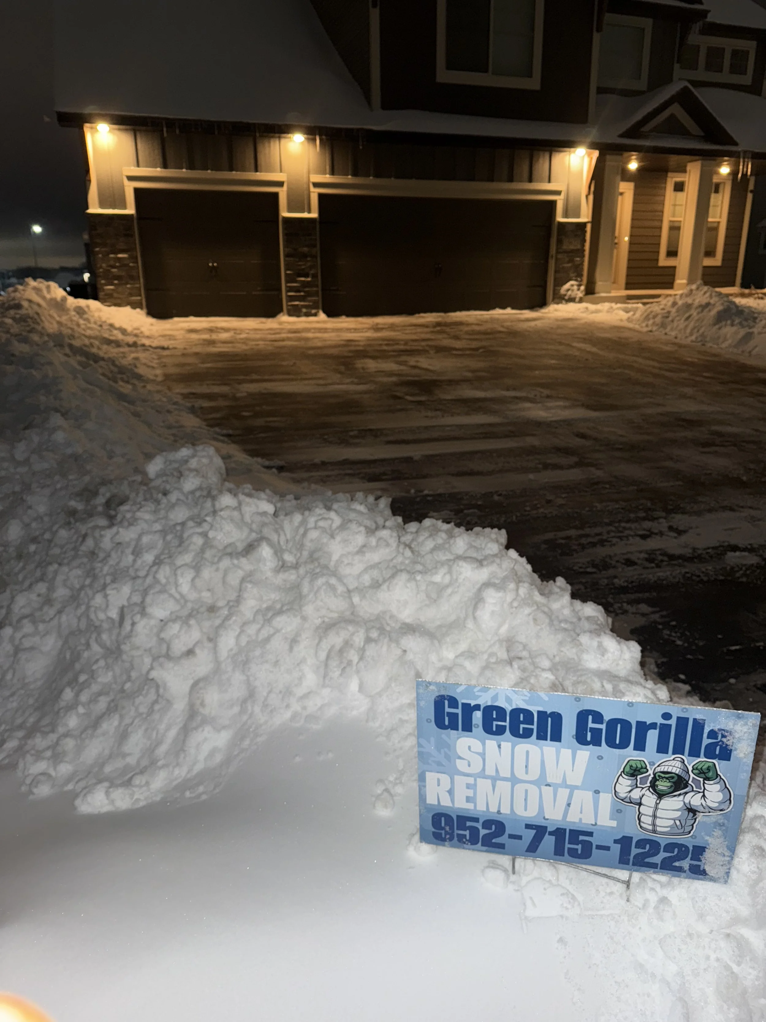 A snowy driveway in front of a house at night, with a sign for Green Gorilla Snow Removal in the foreground.