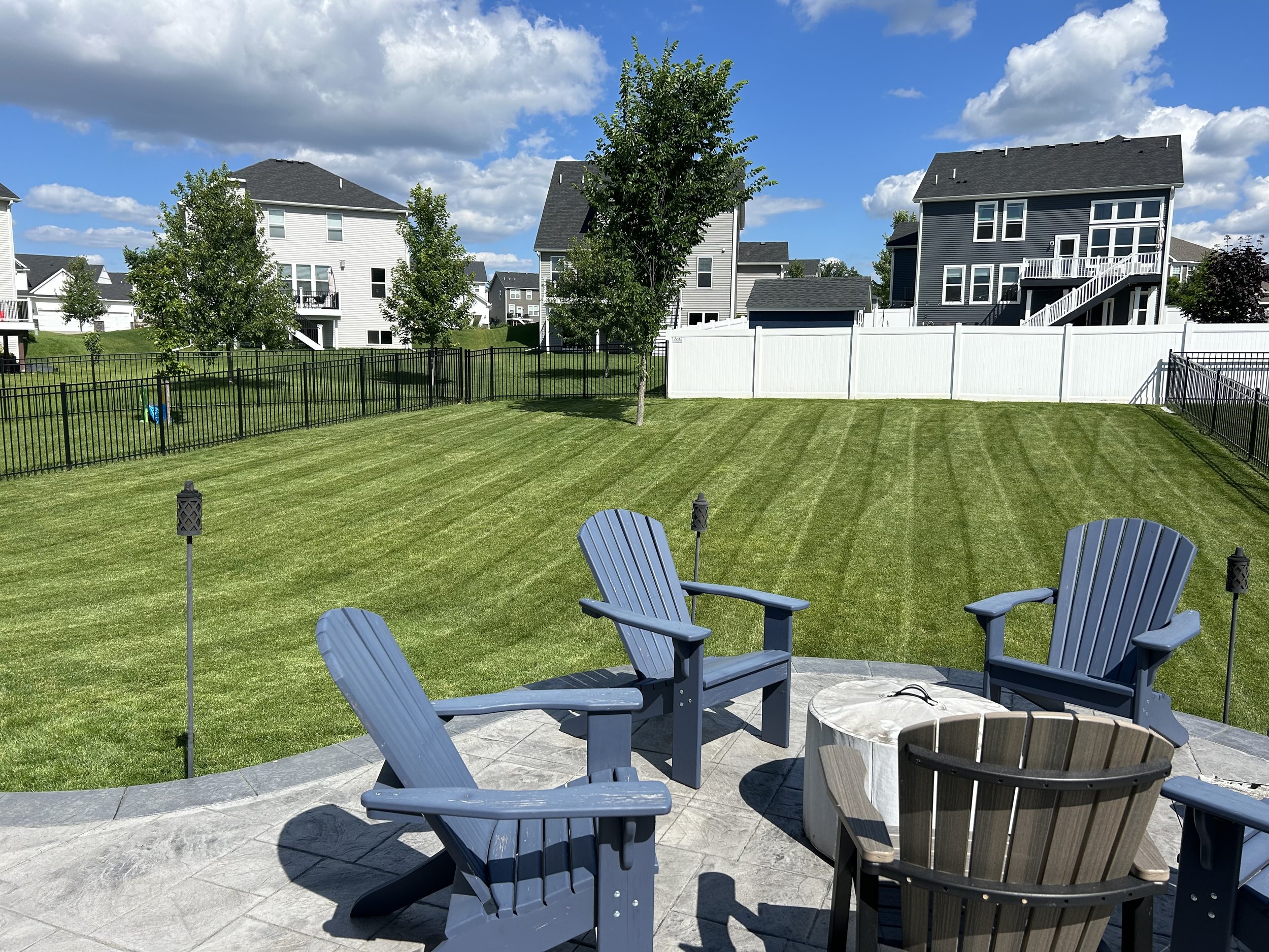 A backyard with a grassy lawn, surrounded by a black and white fence. There are four Adirondack chairs on a stone patio around a fire pit, with a small wooden chair nearby. In the background, there are several modern houses with white and dark gray siding, and a blue sky with some clouds.