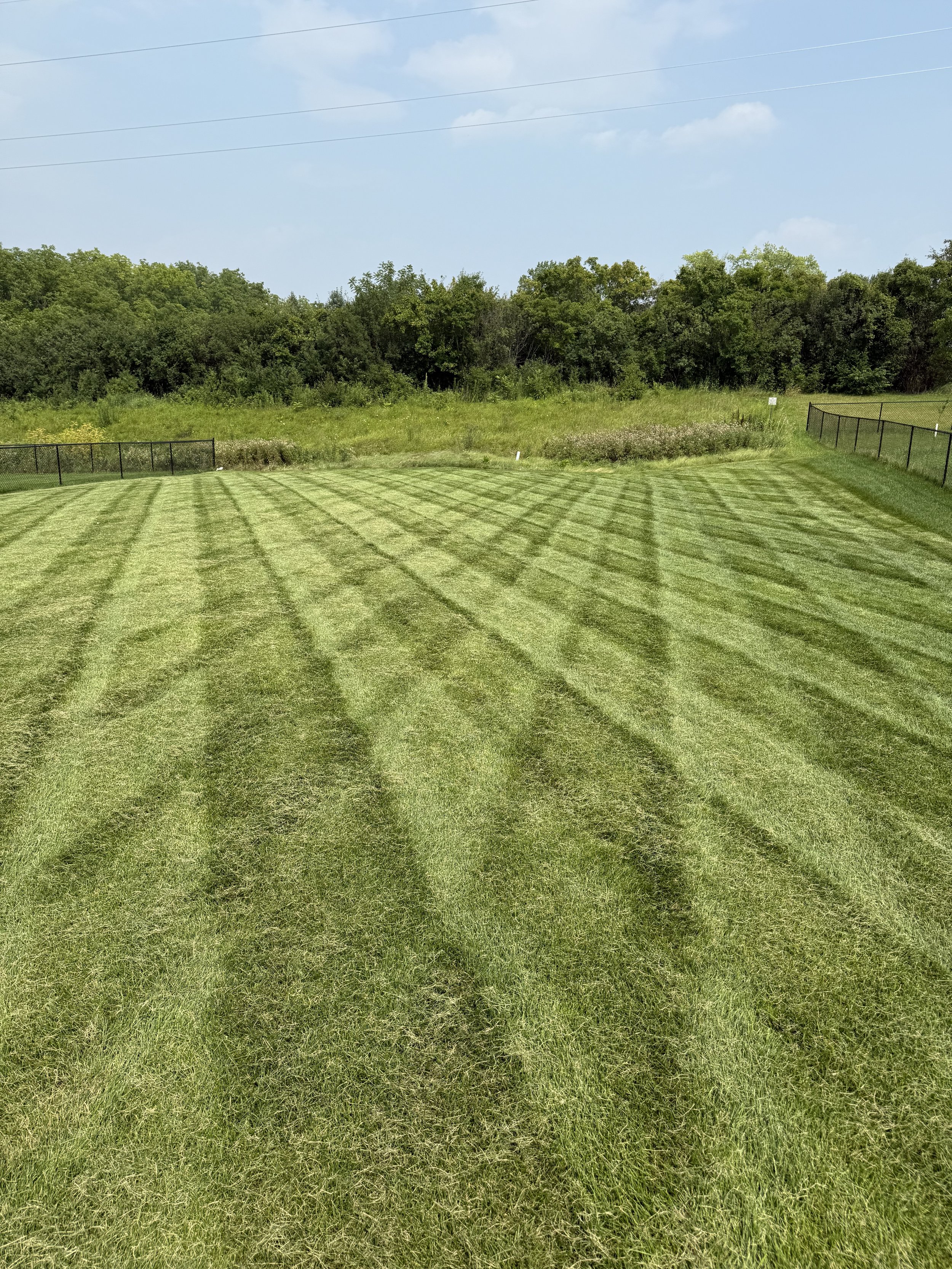 Well-maintained grassy field with freshly mowed pattern, enclosed by a black fence, and bordered by trees and bushes under a partly cloudy sky.