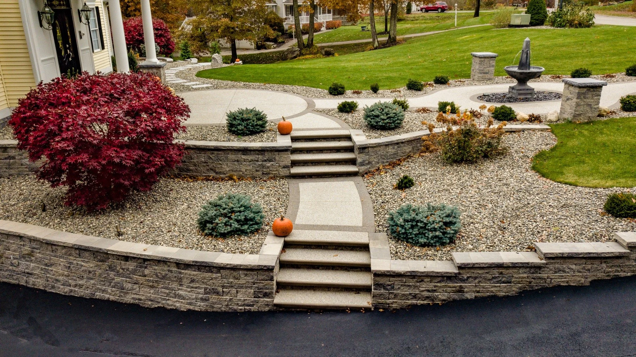 A landscaped front yard with stone retaining walls, stairs, and a pathway, featuring a small fountain, pumpkins, and various bushes, including red and green foliage.