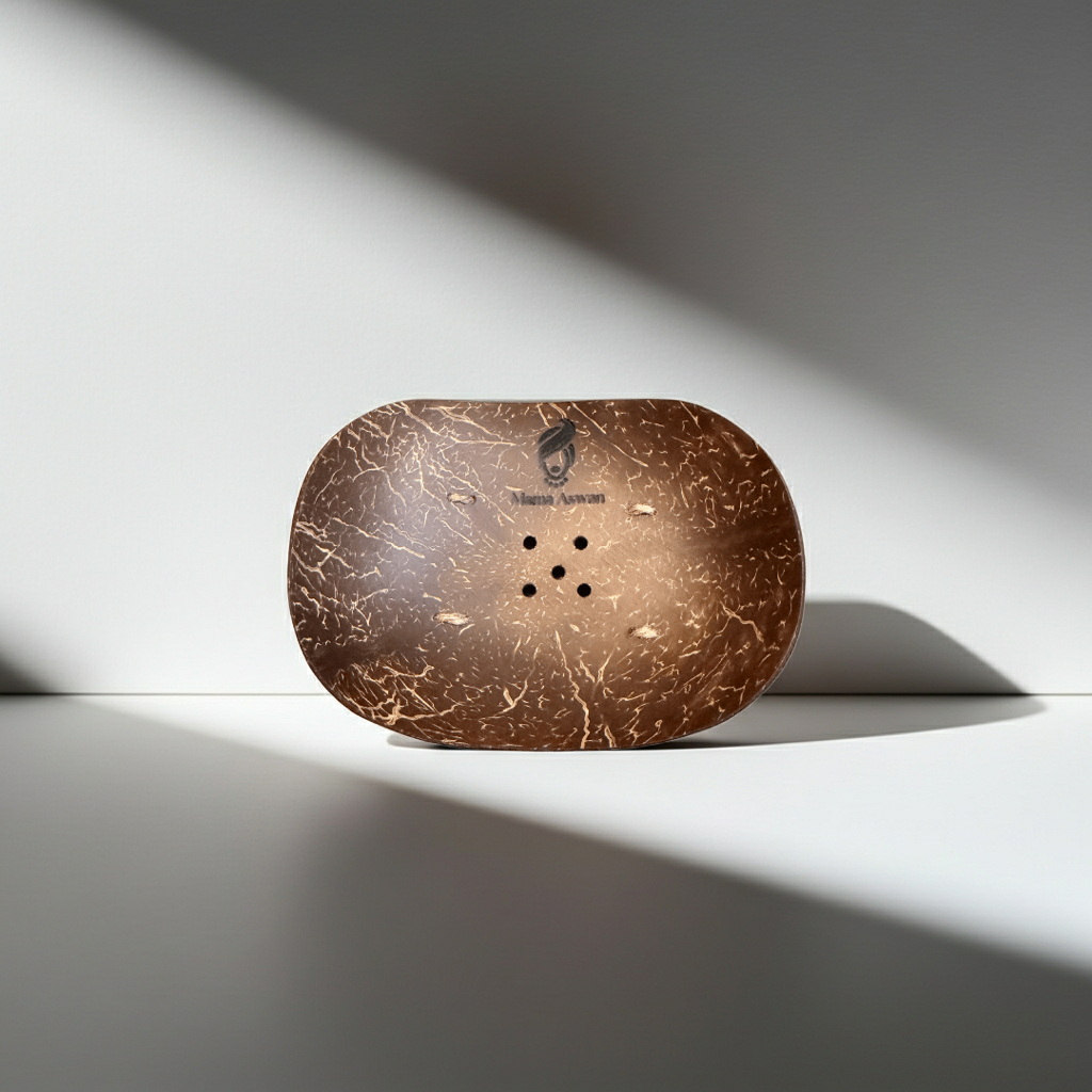 Oval coconut shell soap dish with drainage holes and Mama Aswan logo standing upright in strong natural light.