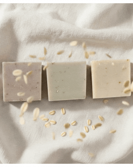 Three oatmeal soap bars arranged on soft cream fabric with scattered oats in warm natural light.
