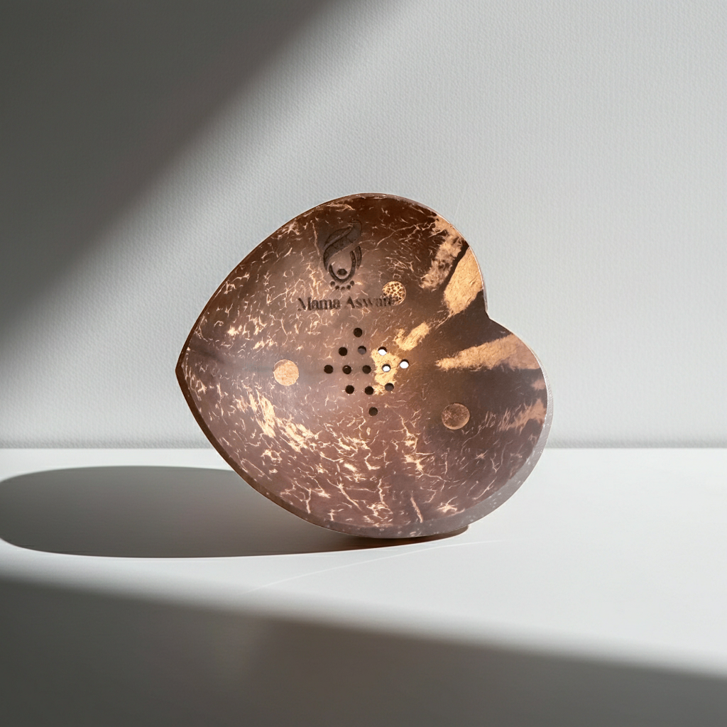 Heart-shaped coconut shell soap dish standing upright in bright light, showing the Mama Aswan logo and drainage holes.