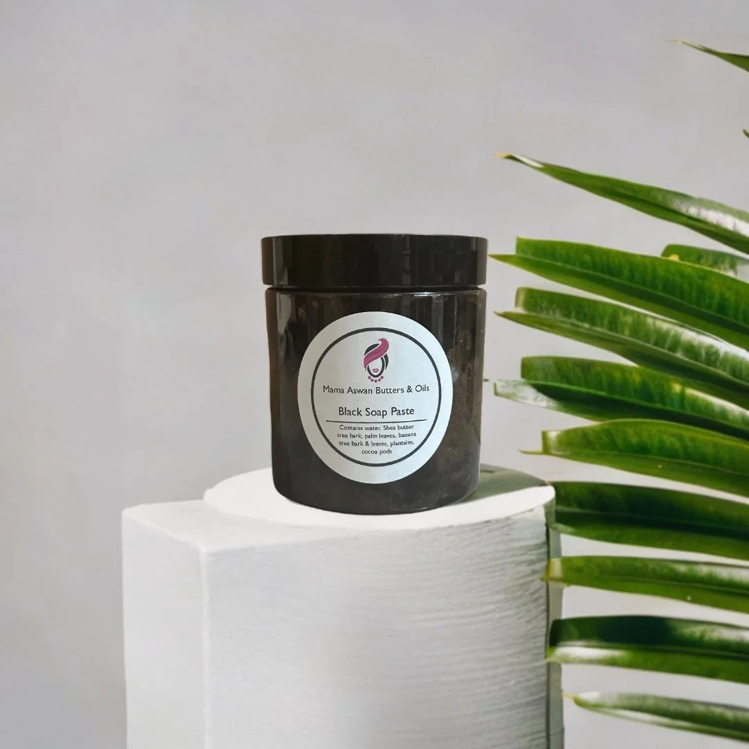 Raw African Black Soap Paste displayed on a white pedestal beside green tropical leaves.