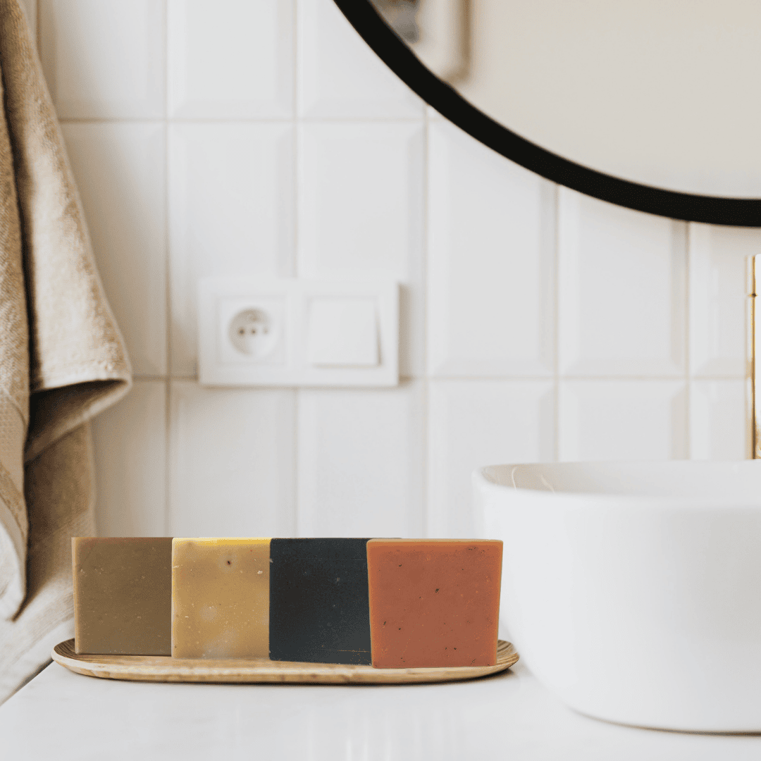 Elegant bathroom countertop with four earthy-toned handmade soap bars in olive green, honey gold, charcoal black, and terra cotta—crafted by Mama Aswan for deep cleansing and daily luxury