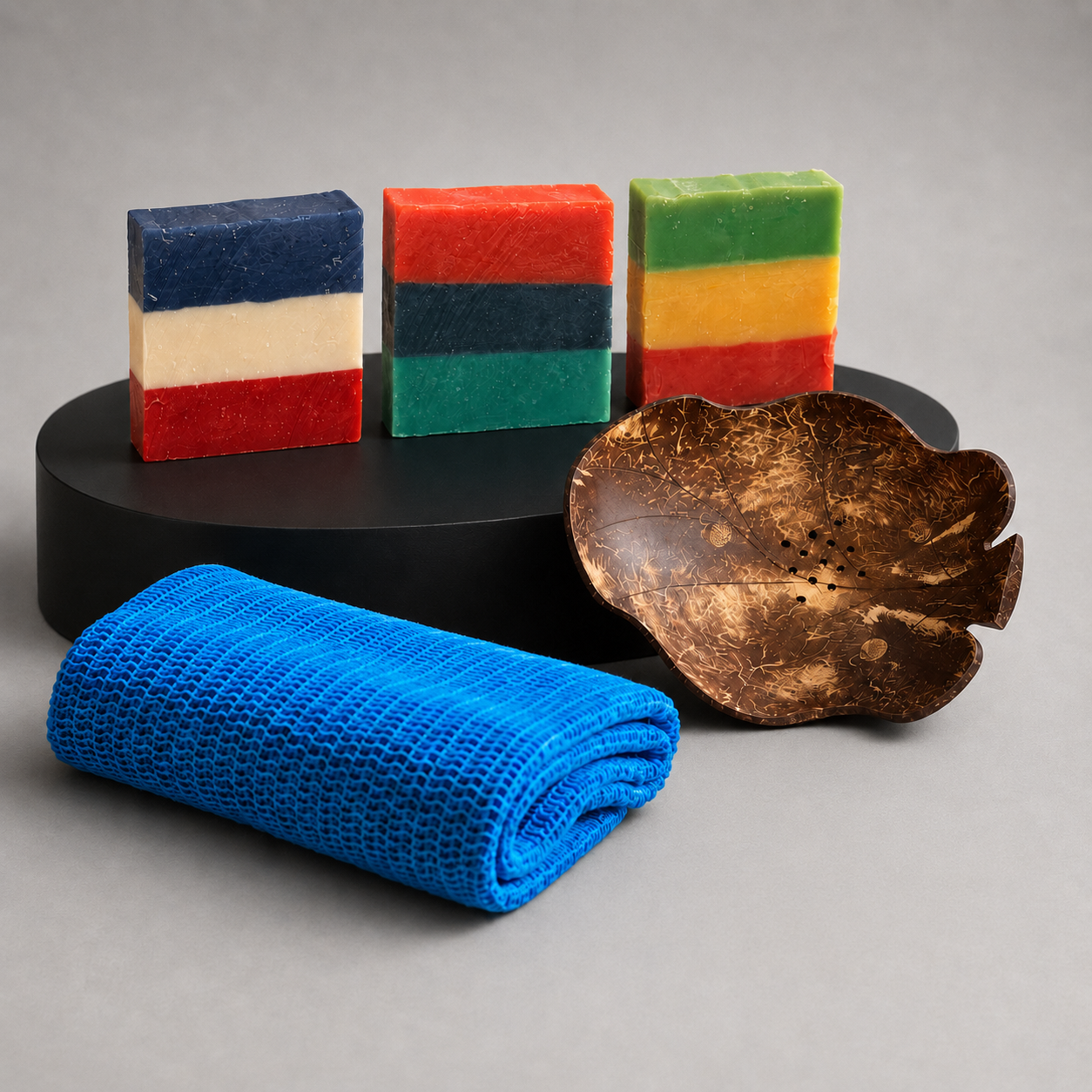 The Heritage Trifecta Shower Set displayed with three Heritage Line soap bars, a blue Sapo Sponge, and a Coconut Shell Soap Dish on a black pedestal.