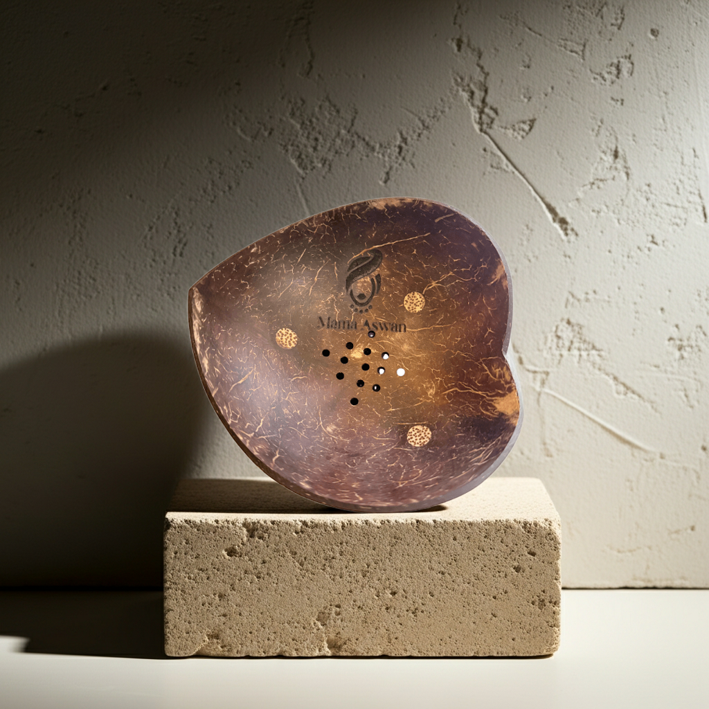 Heart-shaped coconut shell soap dish with Mama Aswan logo displayed on a textured stone block in warm, moody lighting.