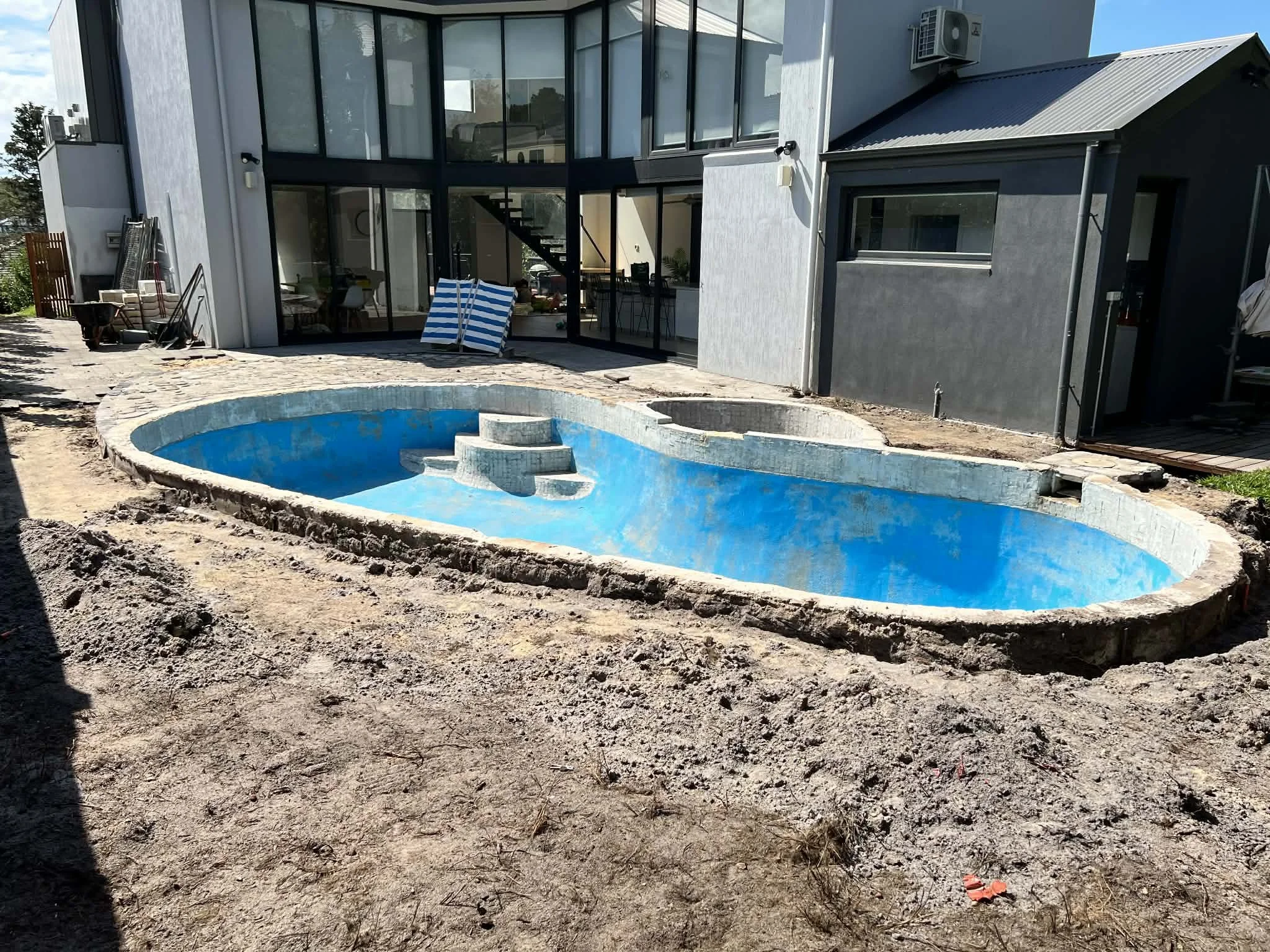 Empty backyard with a pool under construction, house with large windows and patio furniture outside.