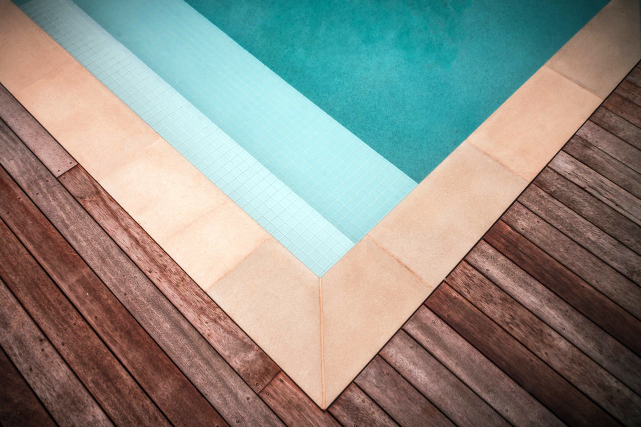 Corner of a swimming pool with light blue tiles and brown wooden deck.