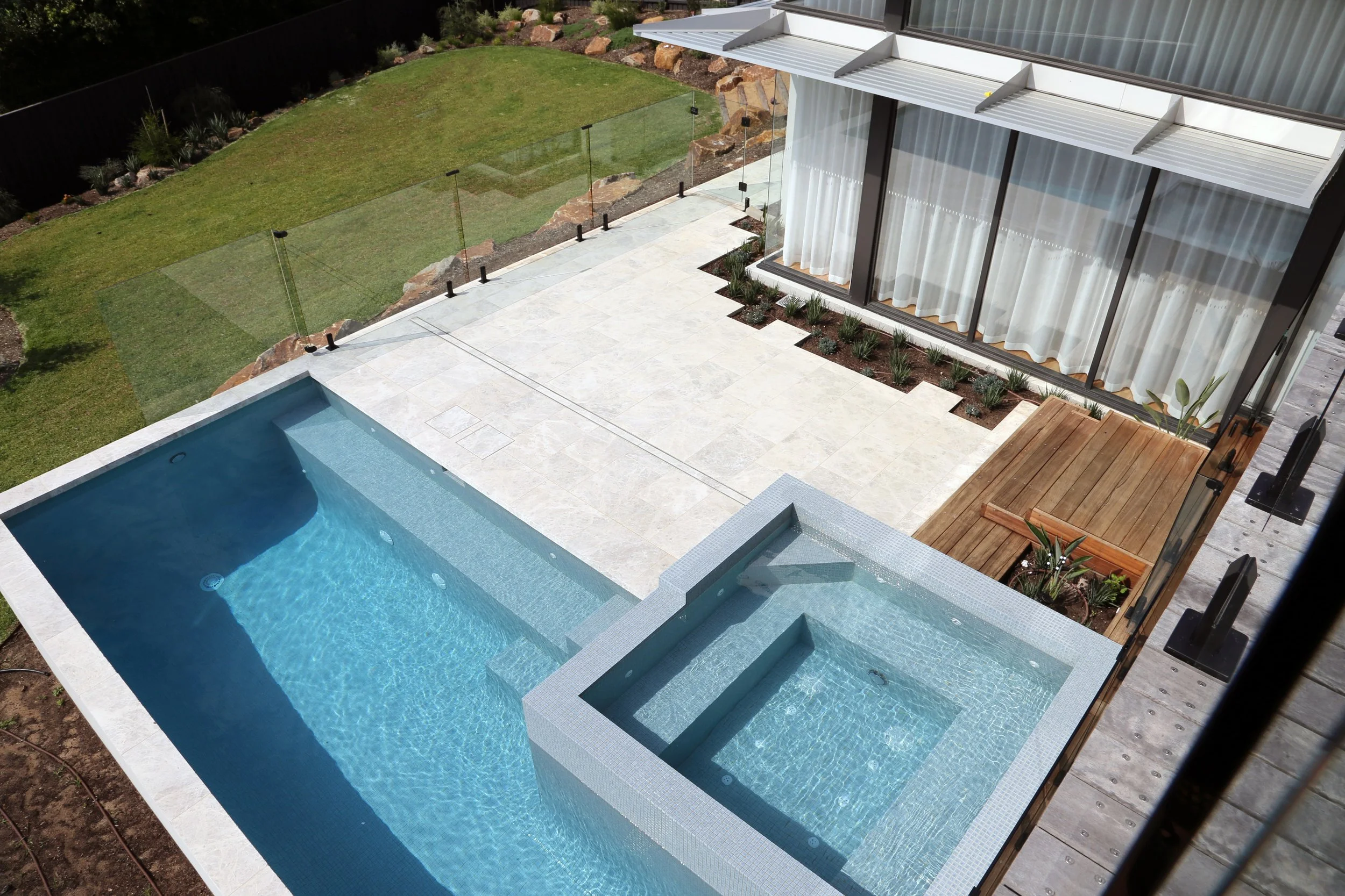 A backyard with a small swimming pool and hot tub, a grassy area with a glass fence, a patio with tiles, potted plants, and a house with large windows and curtains.