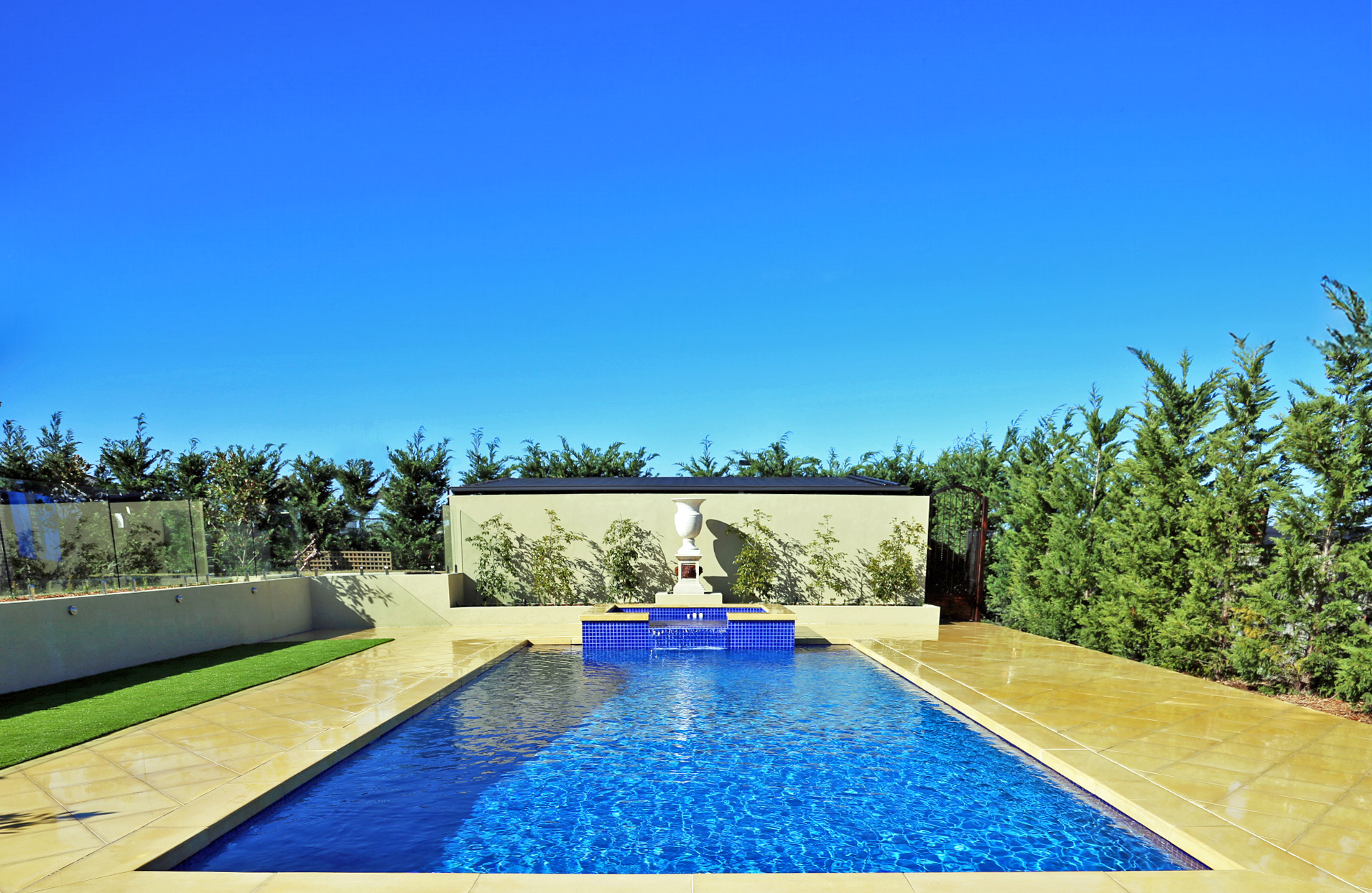 Outdoor swimming pool with blue water, surrounded by a yellow stone deck, green grass, and tall green trees under a clear blue sky.