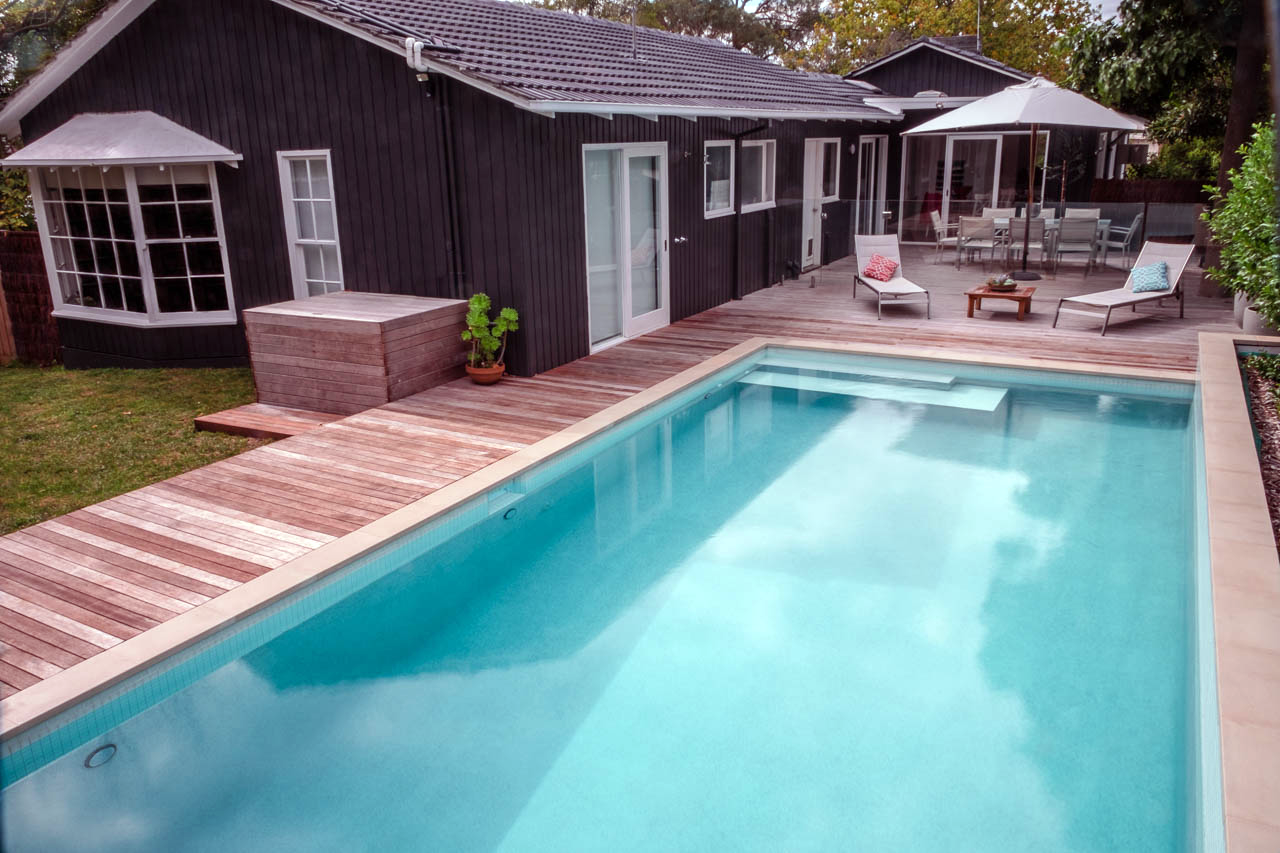 Backyard with swimming pool, wooden deck, patio furniture, umbrella, and house with black exterior