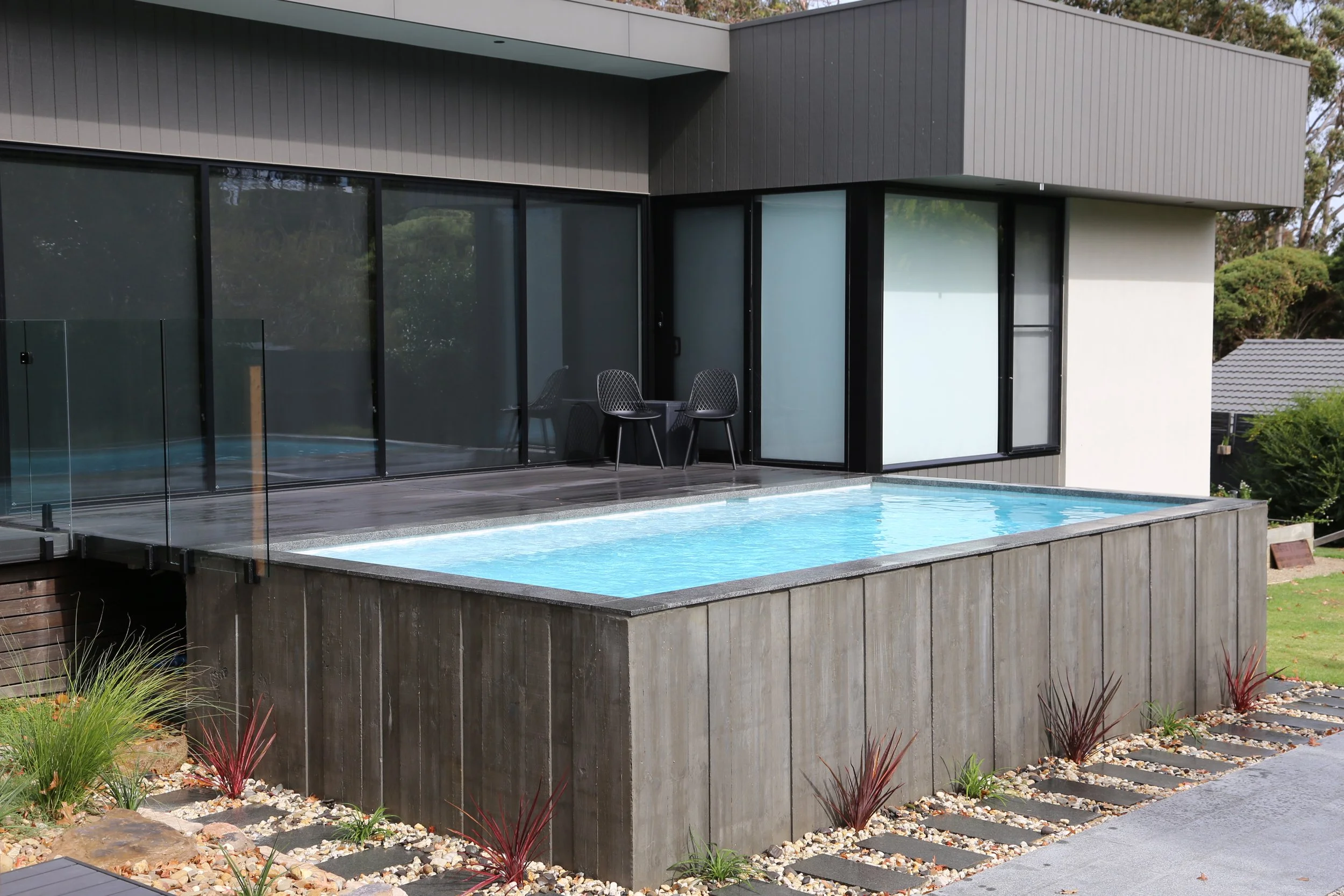 Modern house with glass sliding doors, small above-ground swimming pool on a wooden deck, and landscaped garden with rocks and plants.