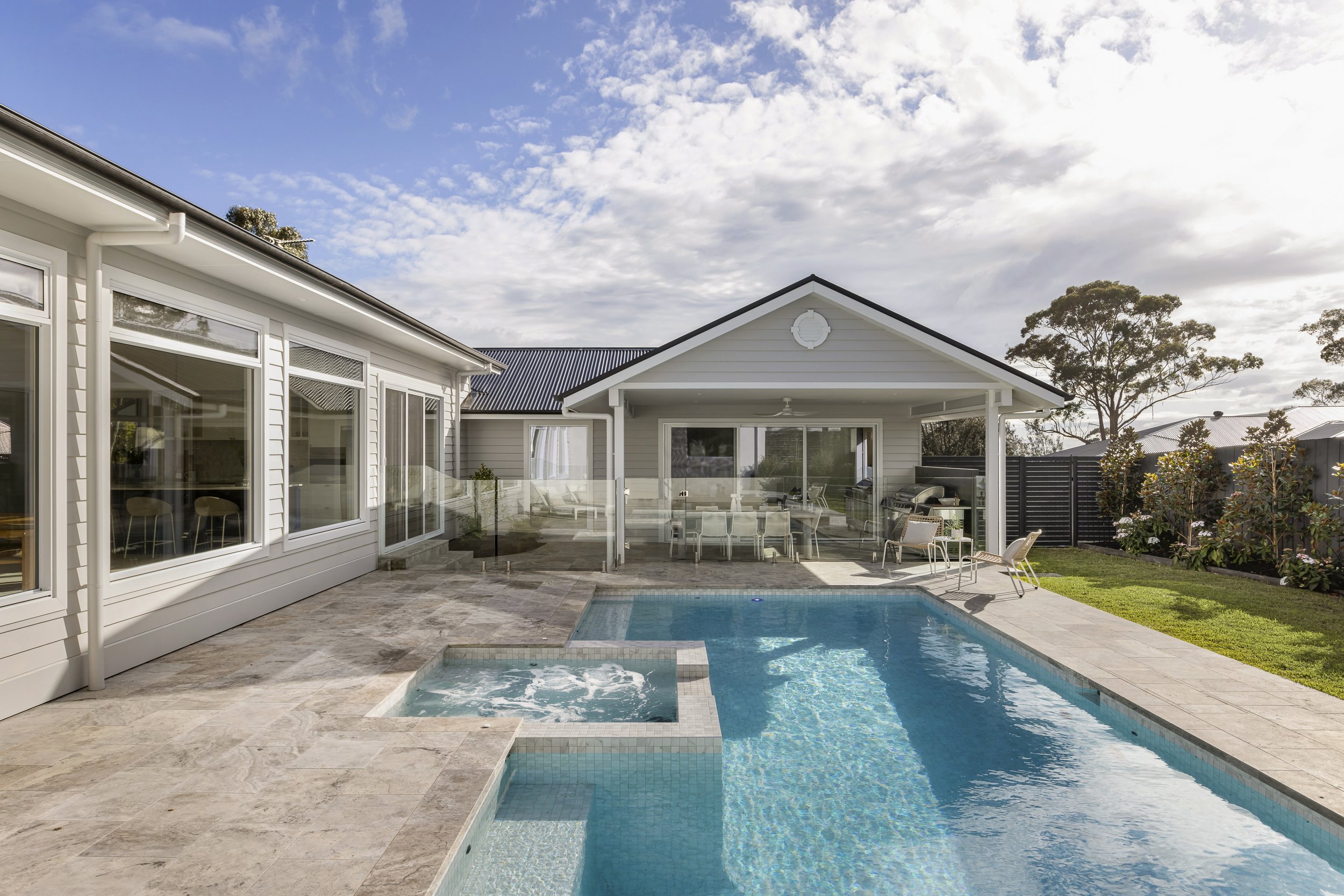 View of the backyard with a swimming pool, patio furniture, and a modern house with large windows under a partly cloudy sky.