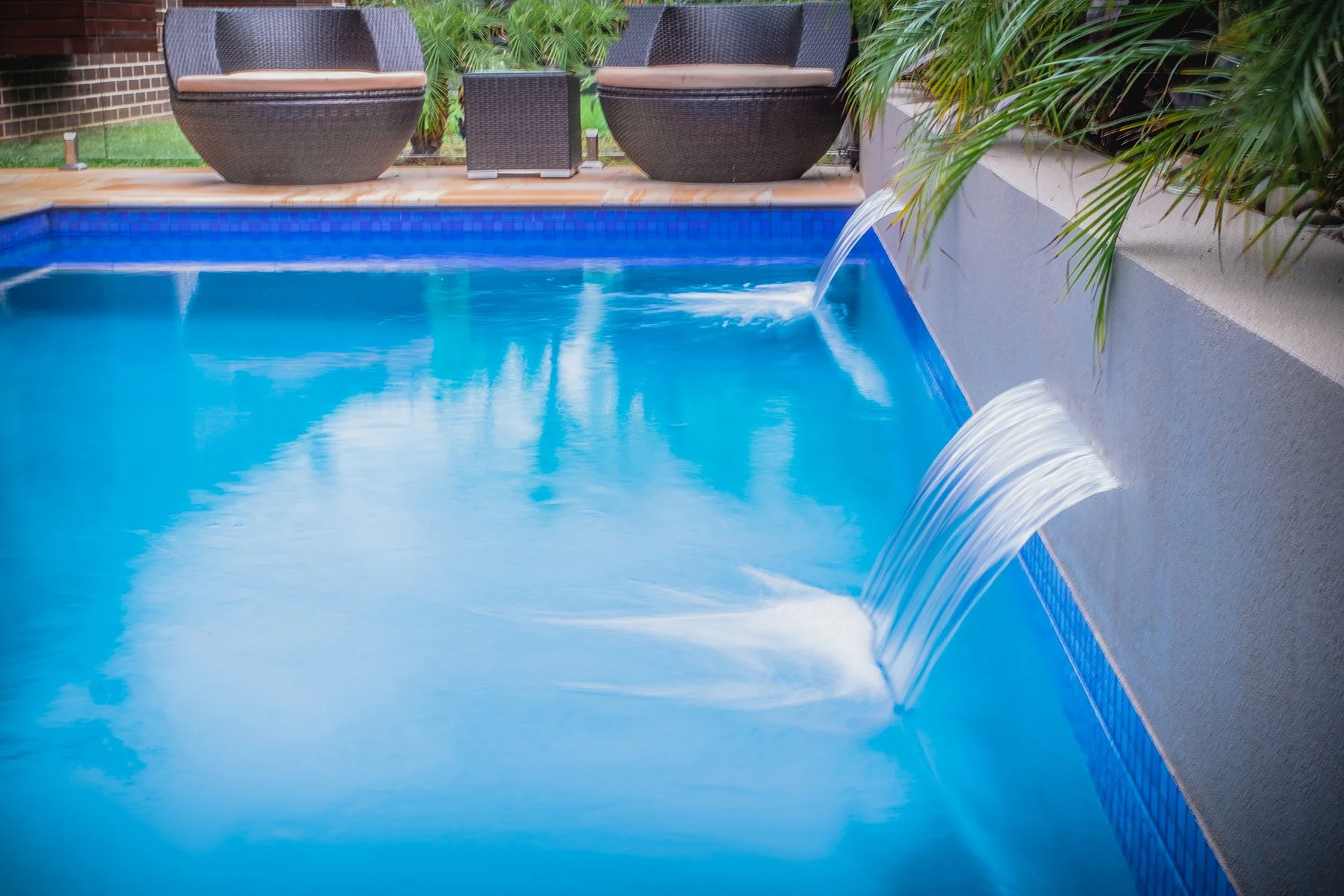 A swimming pool with water features bubbling from spouts on the side, surrounded by patio furniture and green plants.