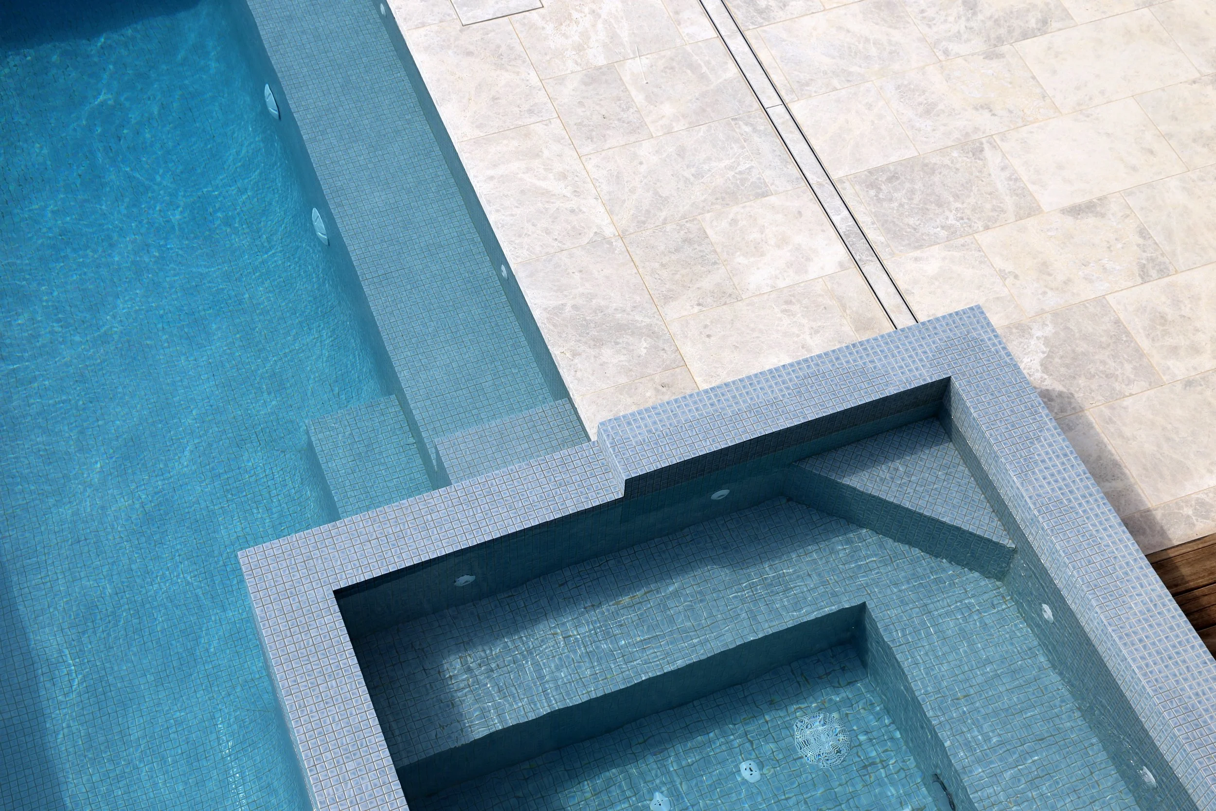 Top view of a modern swimming pool with blue tiled edges and a hot tub with jets, surrounded by beige stone tiles and a wooden deck.