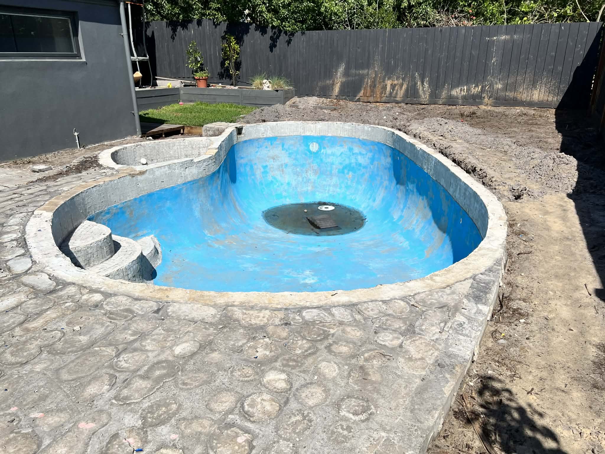 In-ground swimming pool under construction with no water, surrounded by stone patio and a black wooden fence.