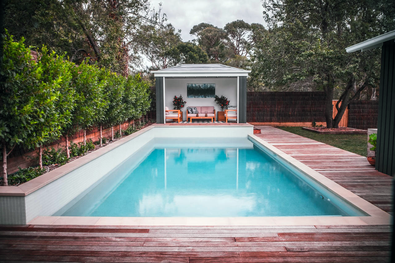 Backyard with a rectangular swimming pool, wooden deck, green shrub wall, and a covered seating area with chairs, sofa, and decorative plants.
