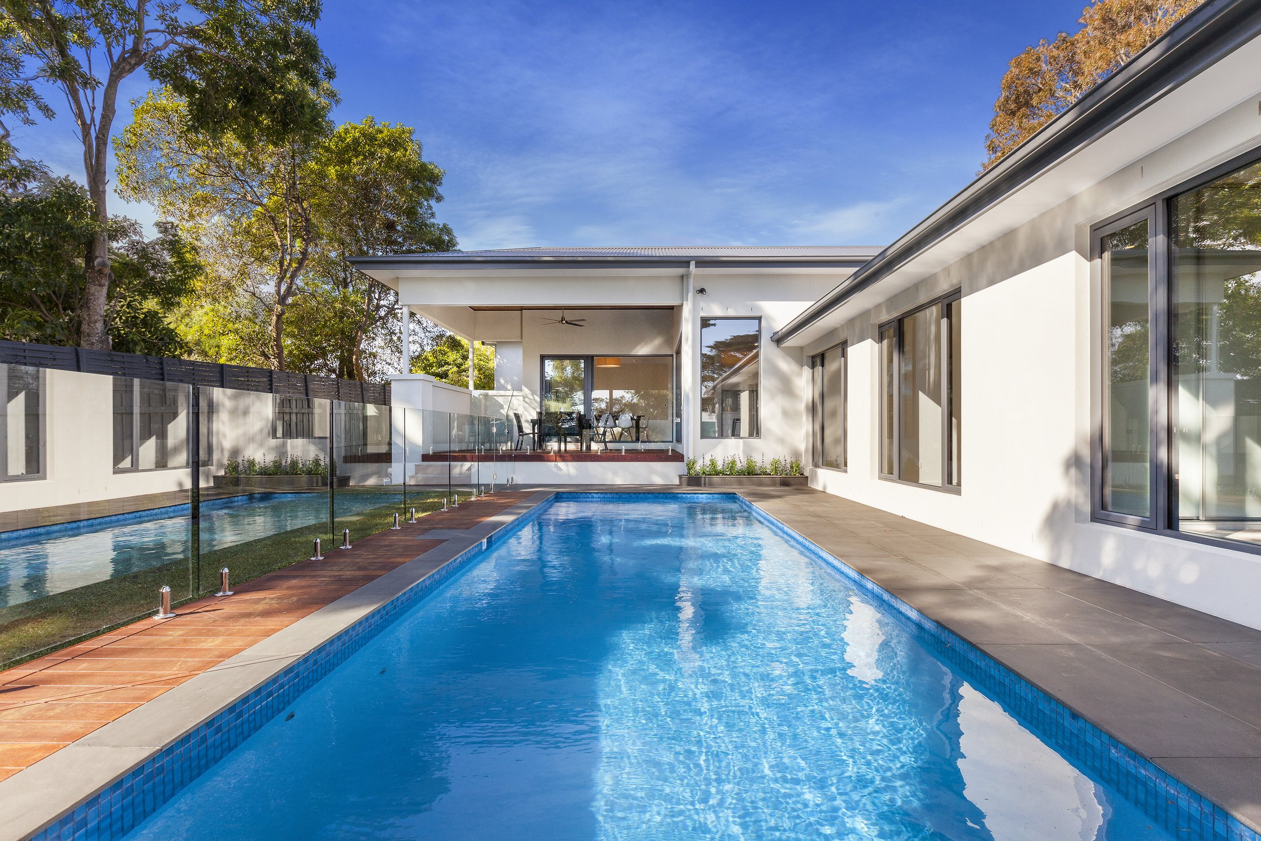 Modern backyard with a blue swimming pool, surrounded by a glass fence and white house with large windows, trees in the background, and a clear blue sky.