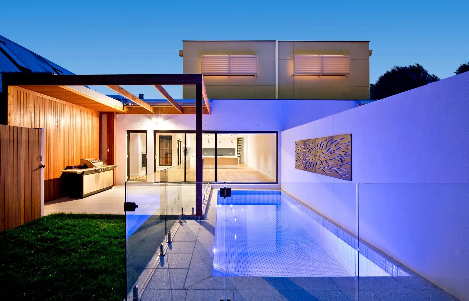 Modern house backyard at dusk featuring a swimming pool with LED lights, glass safety barrier, wooden patio with an outdoor barbecue, and sliding glass doors leading inside.