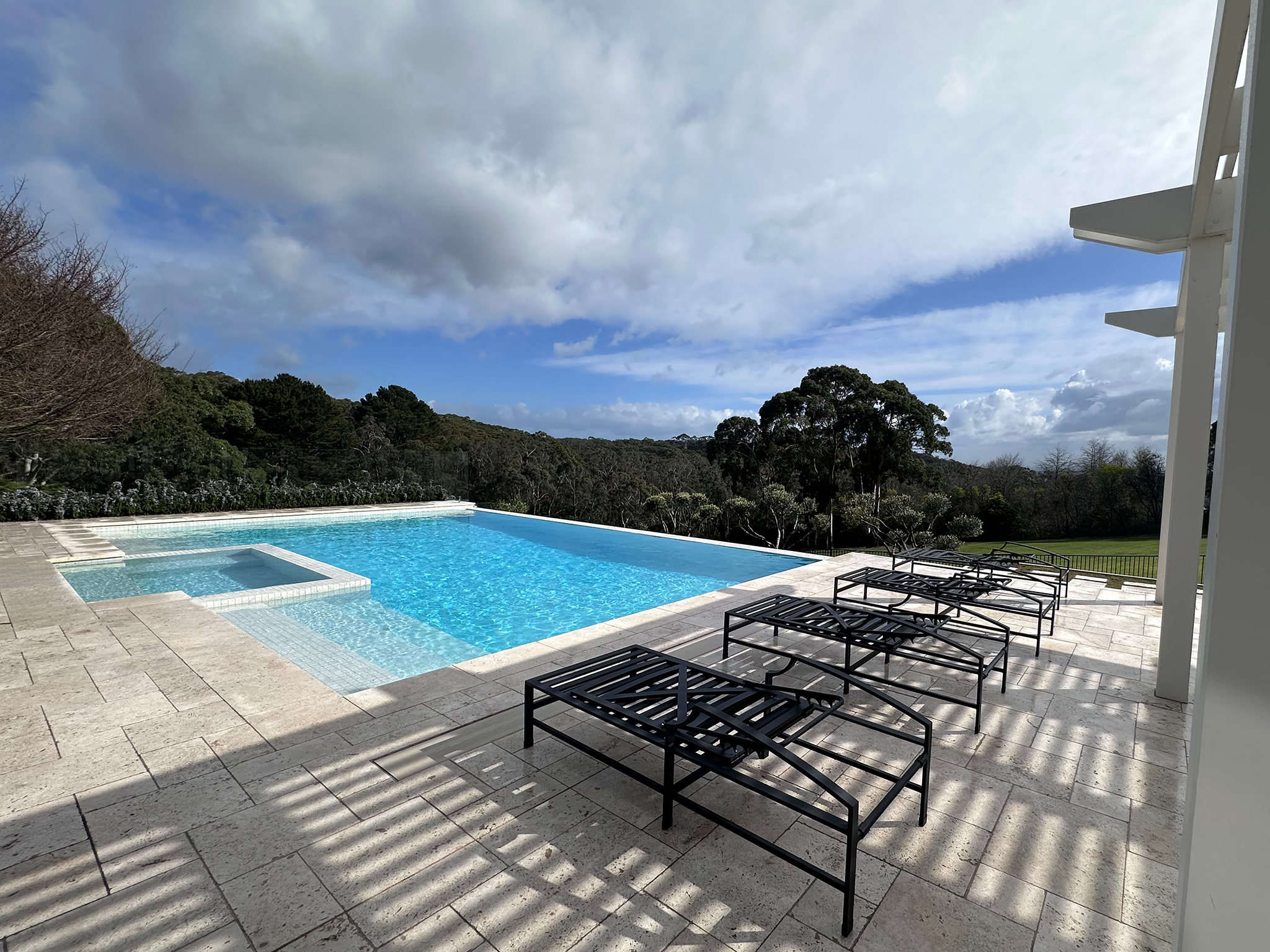 An outdoor swimming pool area with lounge chairs, a view of trees and greenery, and a partly cloudy sky overhead.