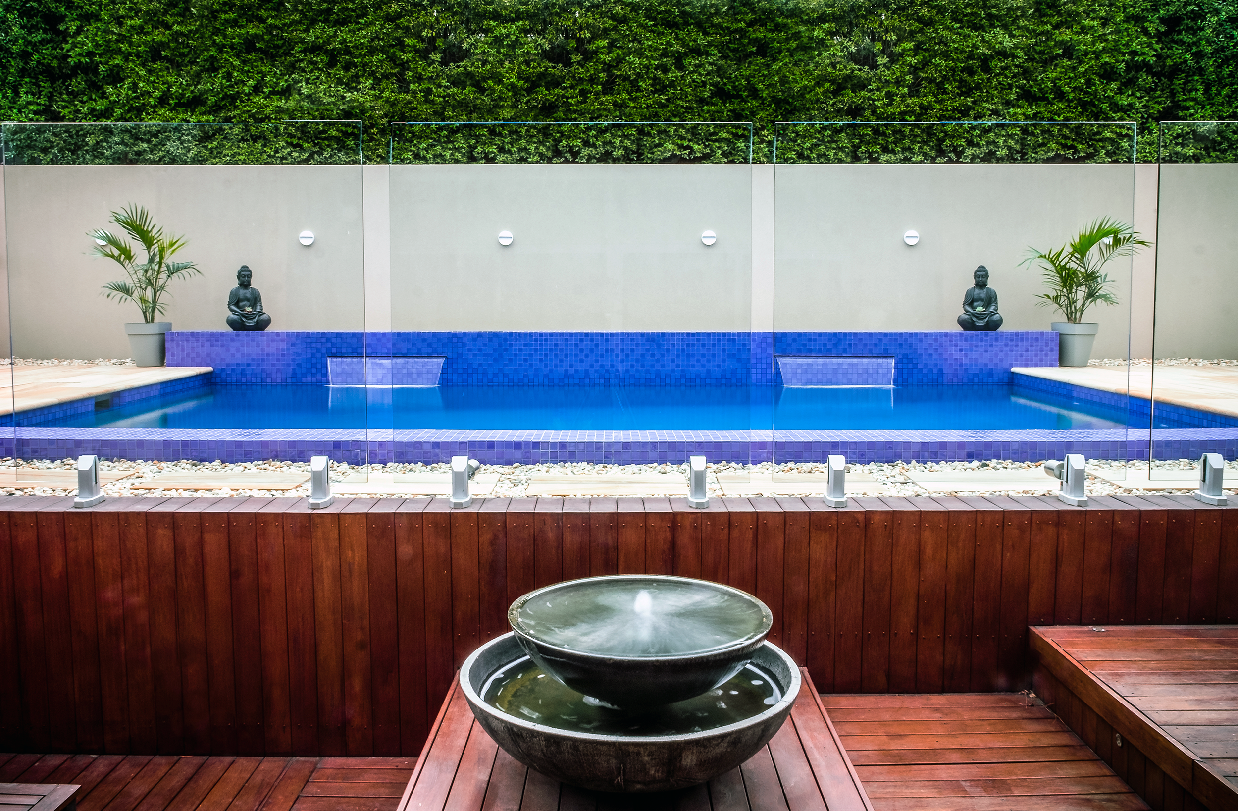 A backyard pool area with a blue-tiled pool, two Buddha statues, potted palm trees, and a privacy fence with greenery on top. In the foreground, there are two black water basins on a wooden deck.