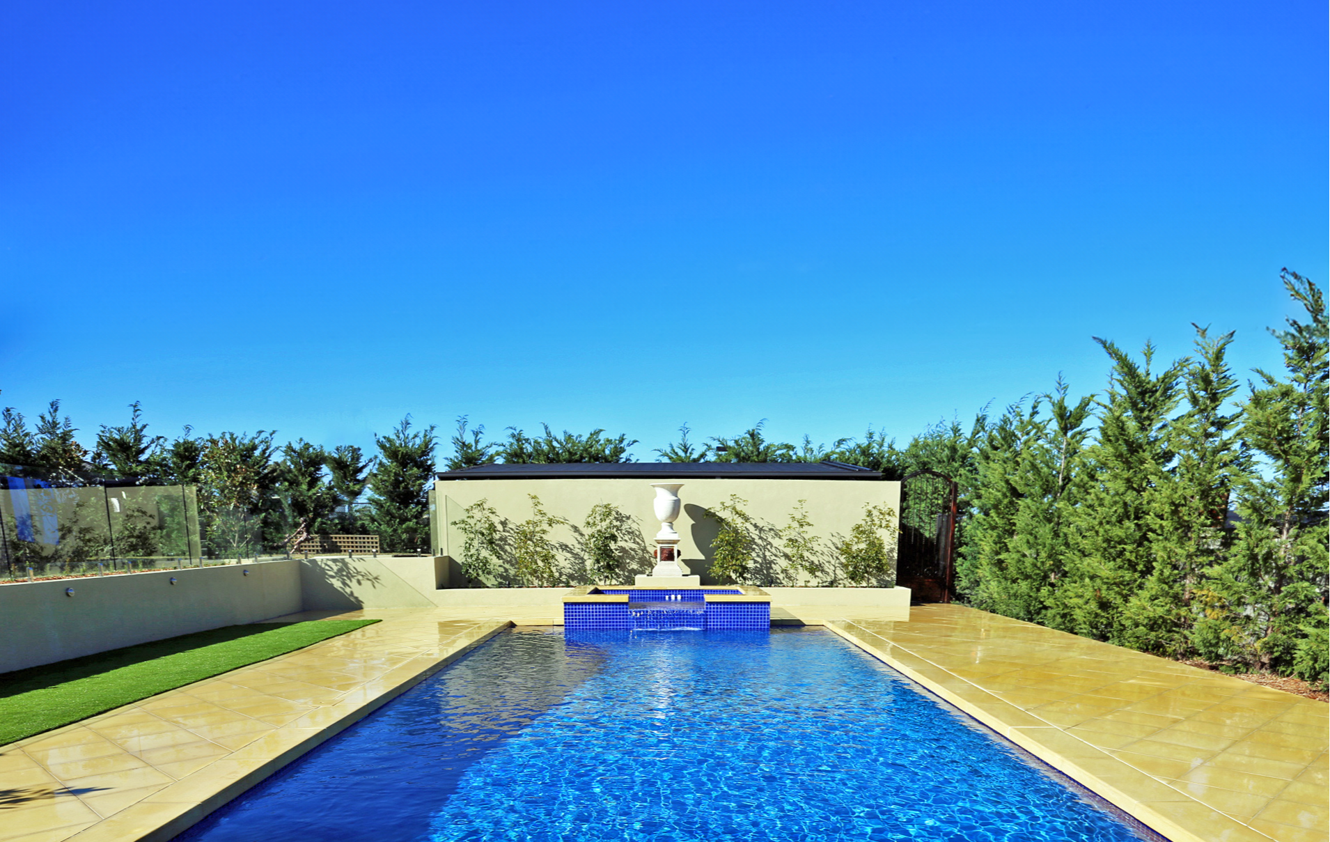 A backyard swimming pool with blue tiles, surrounded by a beige patio and a lawn area. There is a decorative feature with a white urn on a pedestal in front of a white wall, and lush green trees and bushes in the background under a clear blue sky.