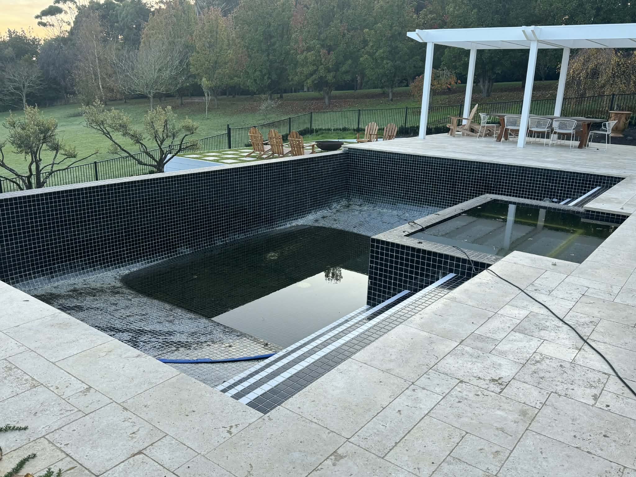 An outdoor backyard area with a pool that is under construction or renovation. The pool has black tiles and the surrounding area is paved with large light-colored tiles. There are several wooden chairs and a table with white chairs on a patio area underneath a white pergola. In the background, there is a green lawn with trees and a black fence.