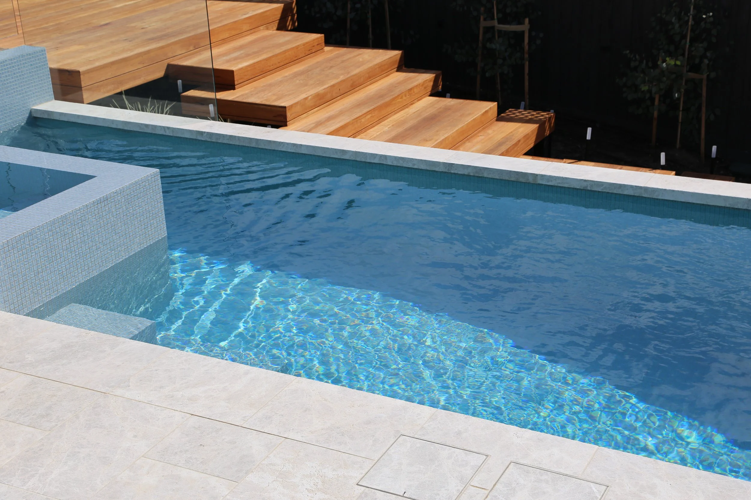 Side view of a luxurious pool in Mount Martha on the Mornington Peninsula with white pavers, blue water and wooden decking surrounding it.