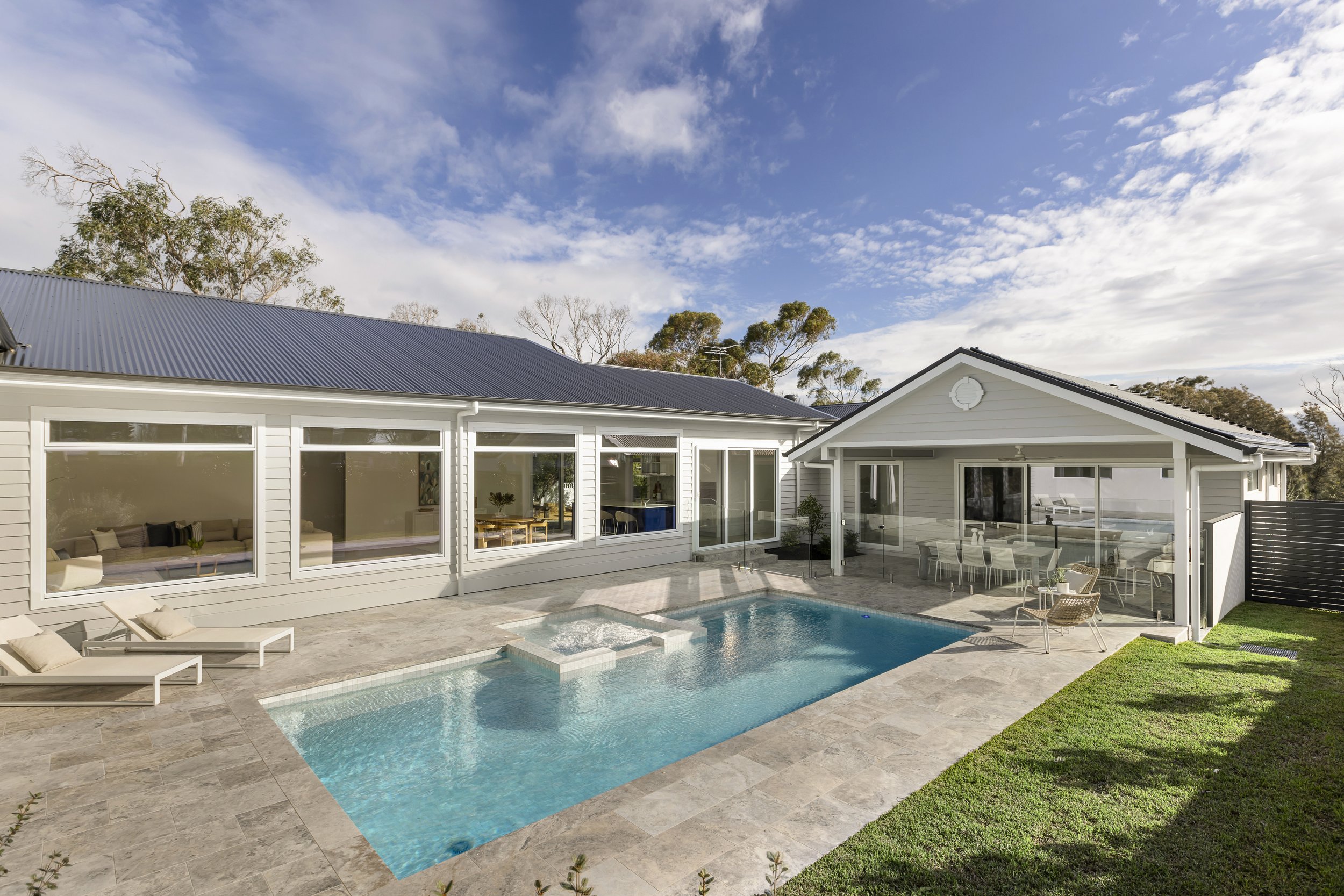 A modern house with large windows, a backyard pool, outdoor seating, and a partly cloudy sky.