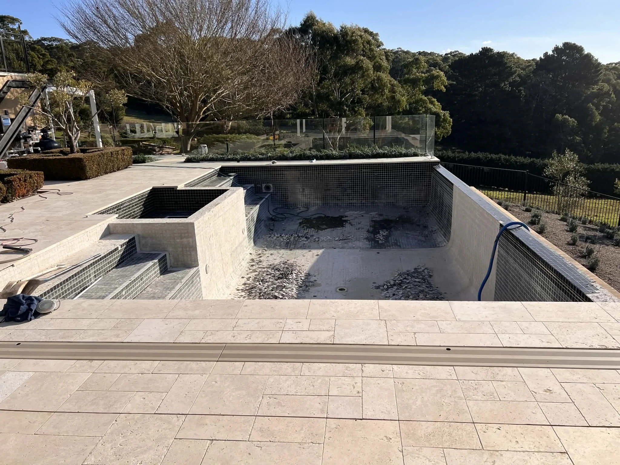 An outdoor scene shows a partly constructed pool with tile lining, surrounded by a spacious patio with beige tiles. There are trees, shrubs, and a hillside with dense greenery in the background. Construction tools and debris are visible around the pool area.