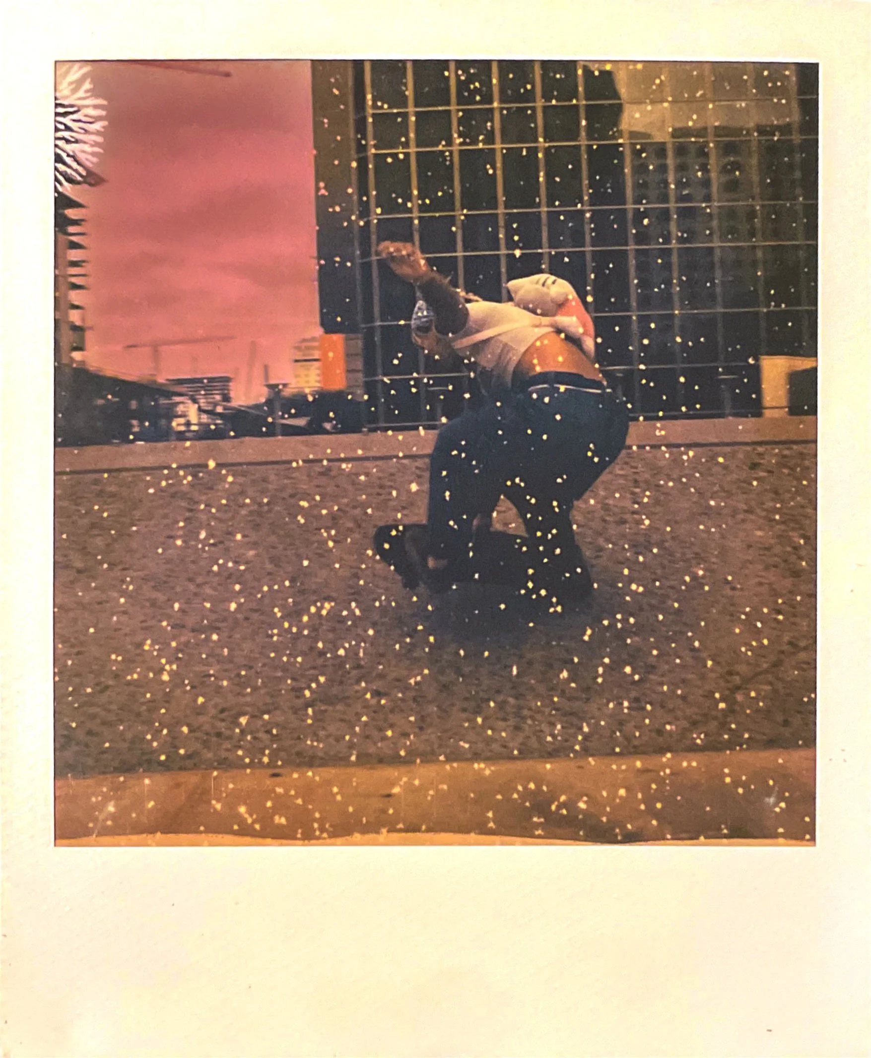 A person dancing or performing in front of a cityscape with tall buildings and a cloudy sky, during dusk or nighttime, with a visual overlay of golden sparks or small particles.
