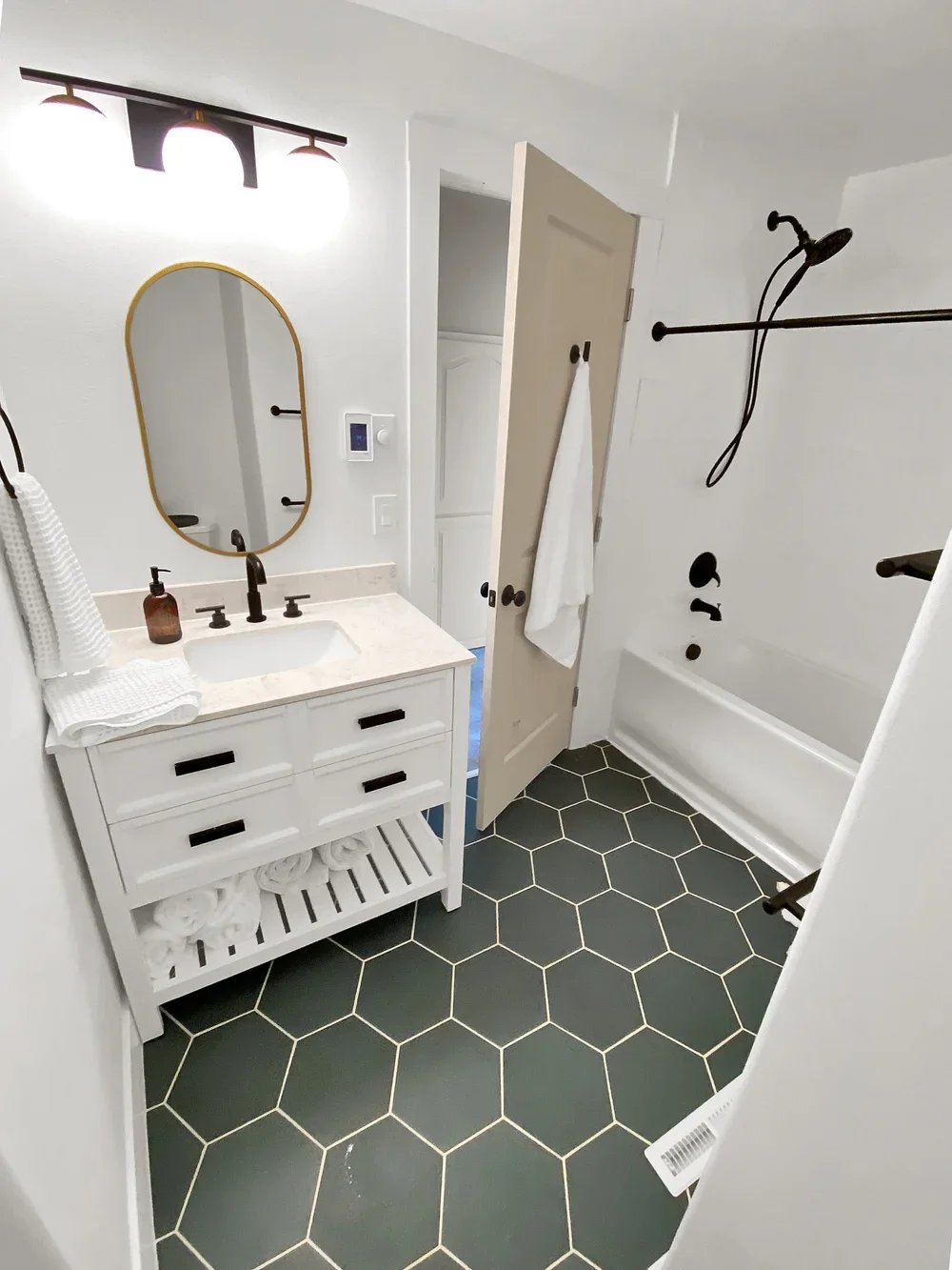 A modern bathroom with a white vanity, oval mirror with a gold frame, black hardware, and black hexagon floor tiles. It features a walk-in shower with black fixtures and a white bathtub with a black showerhead and rod.