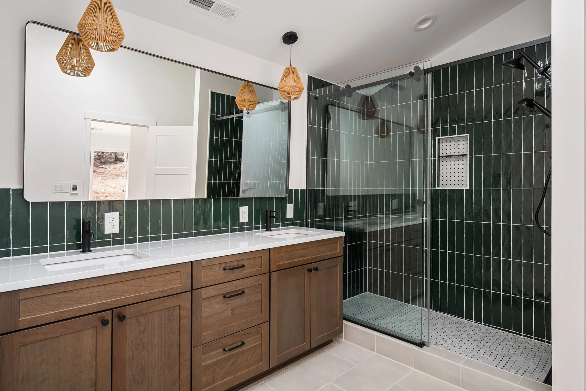 Modern bathroom with double vanity, dark green tile backsplash, large mirror, and walk-in shower with glass door and black fixtures.