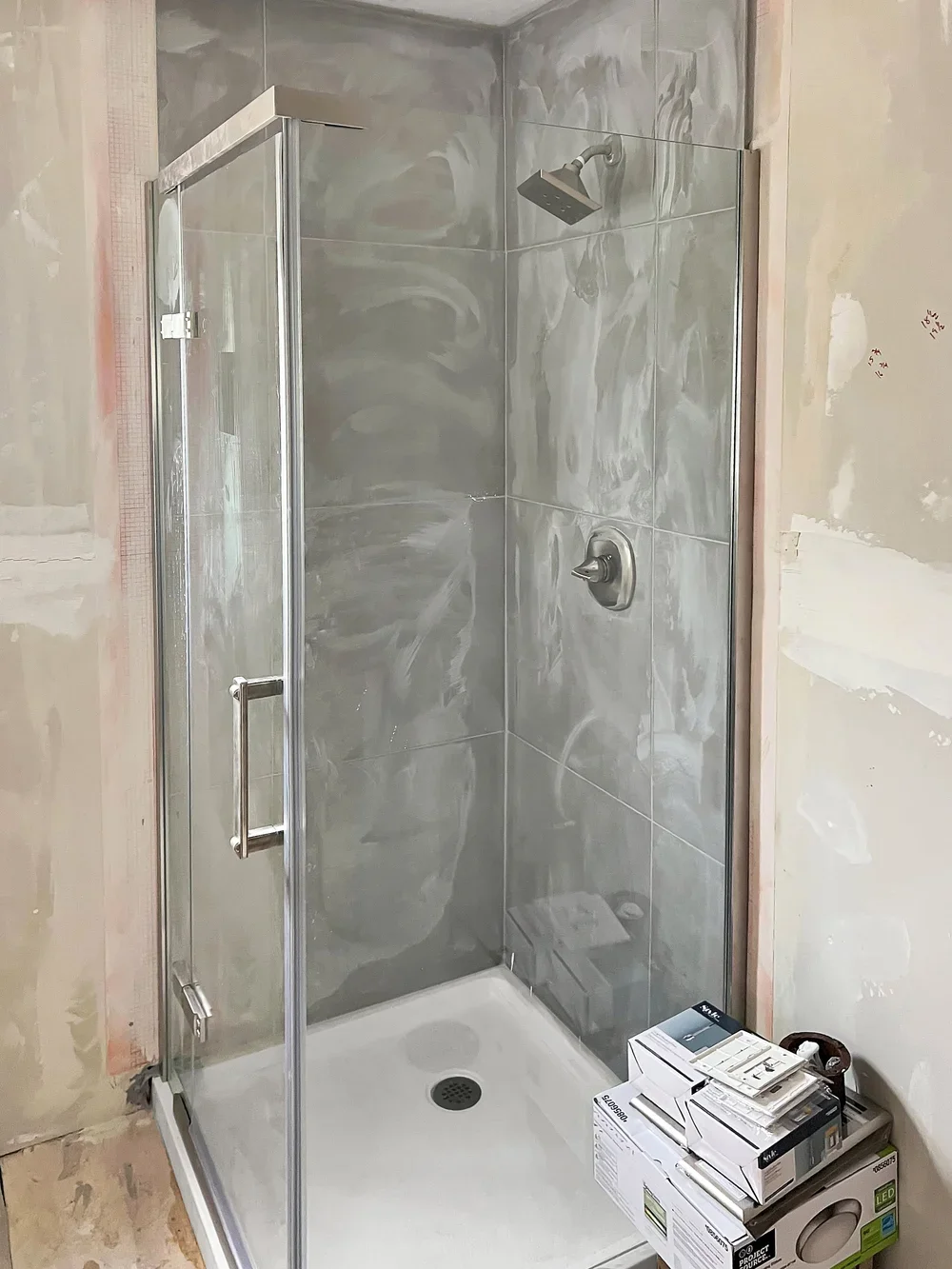 A newly installed glass shower enclosure with metal framing, a square showerhead, and a single handle for water control, located in a bathroom under construction or renovation.