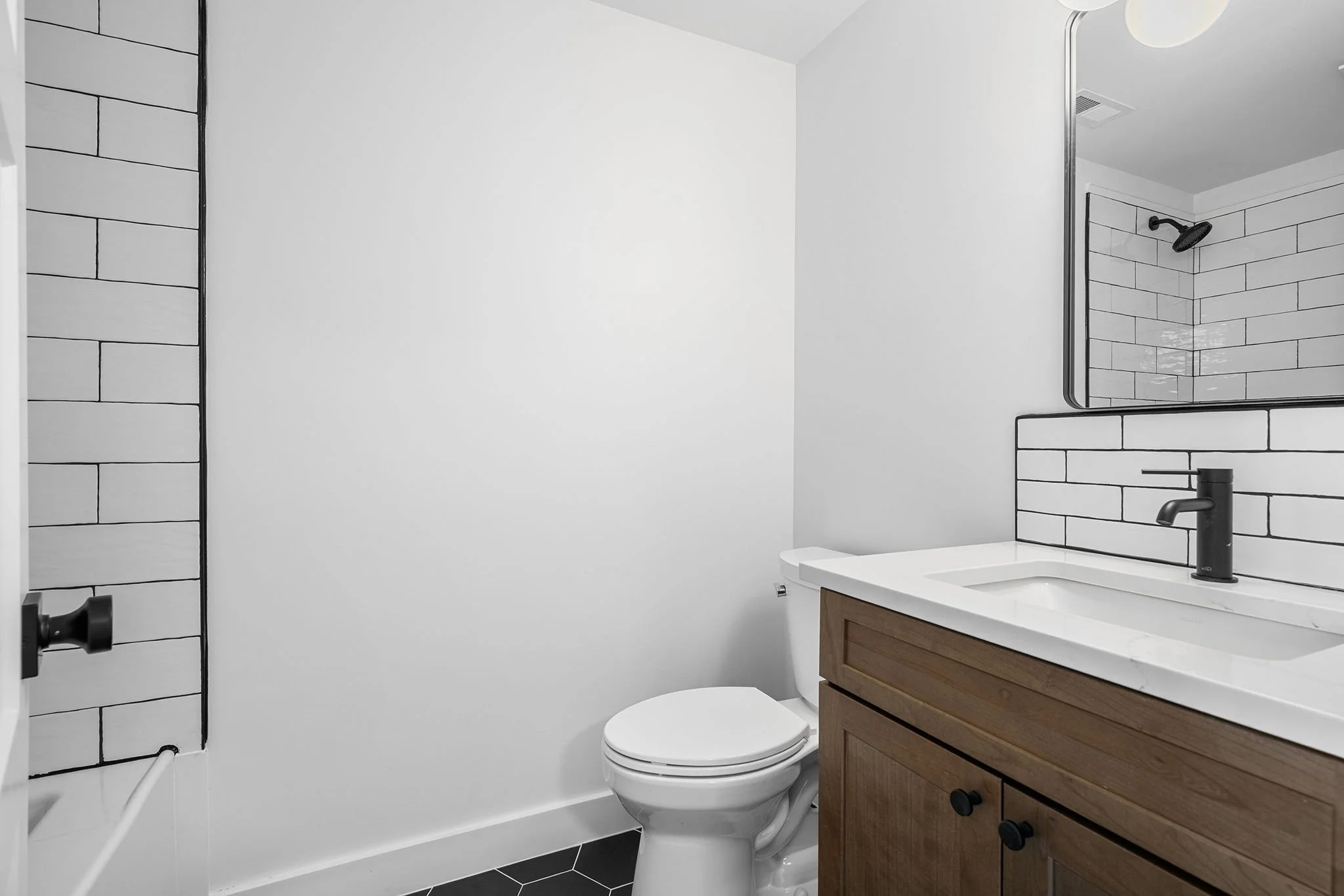 Modern bathroom with white walls, a wooden vanity with a white sink, a black faucet, a mirror, black subway tile backsplash, and a toilet.