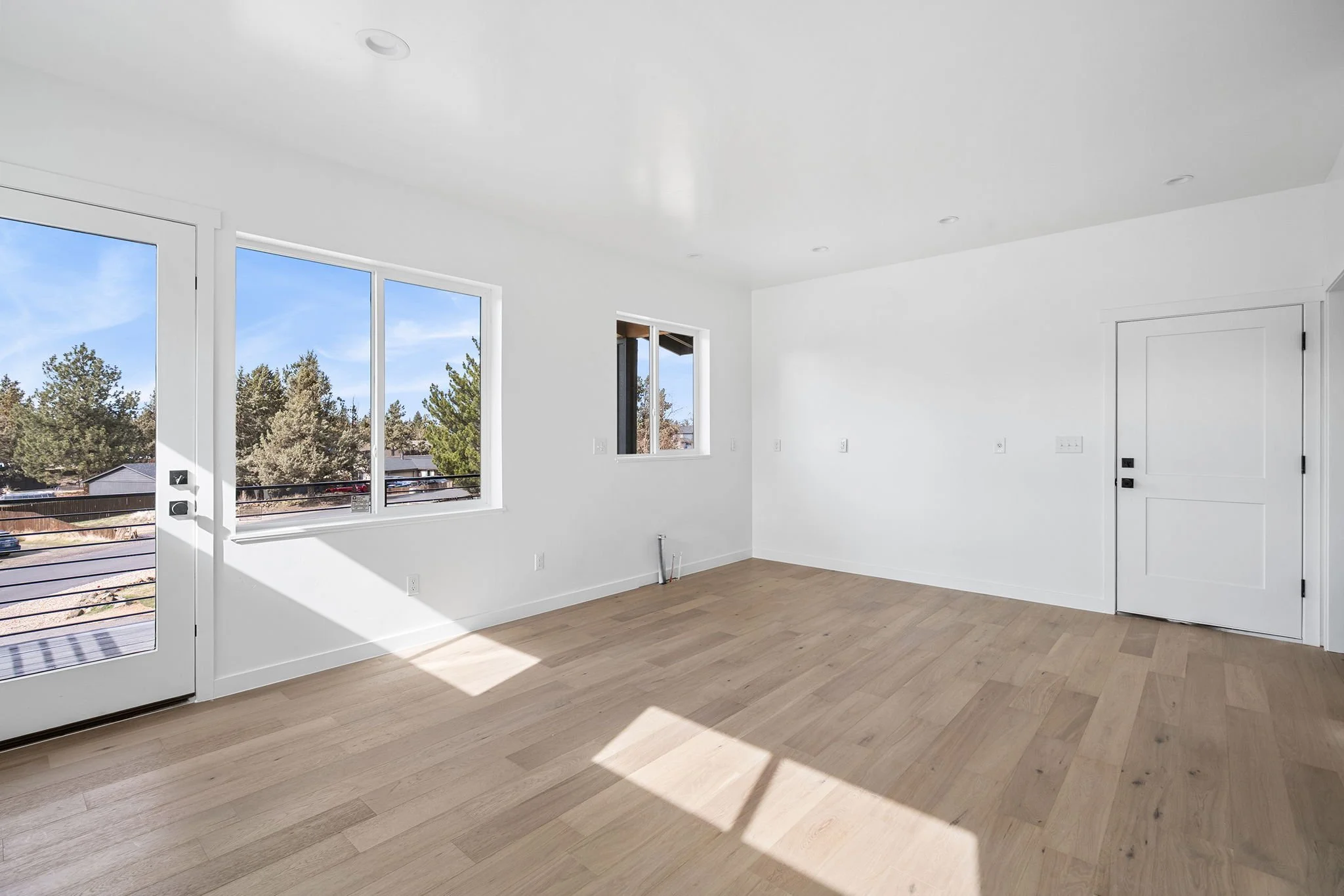 Empty living room with large windows, white walls, light-colored wooden floors, and a door leading outside.