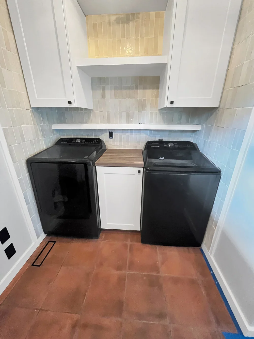 Laundry room with two black washing machines, white cabinetry, beige tiled walls, and brown tiled floor.