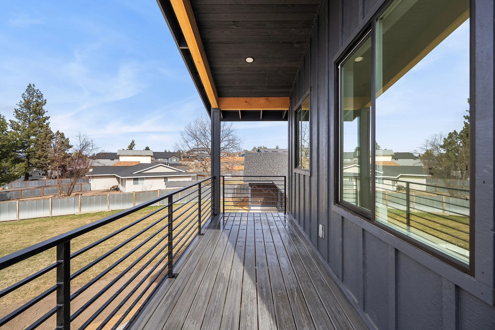 View of a modern house balcony with wooden flooring, black metal railing, and large windows on the side, overlooking a backyard with a wooden fence and neighboring houses under a clear blue sky.