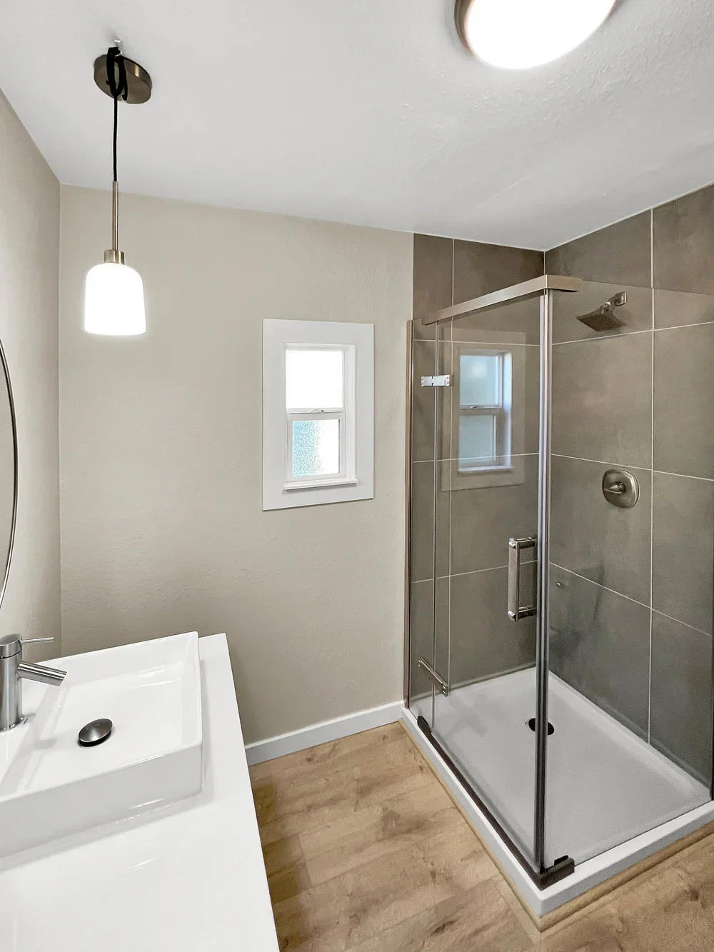 A modern bathroom with a glass shower enclosure, a small window, a white sink with a round mirror, and hardwood flooring.