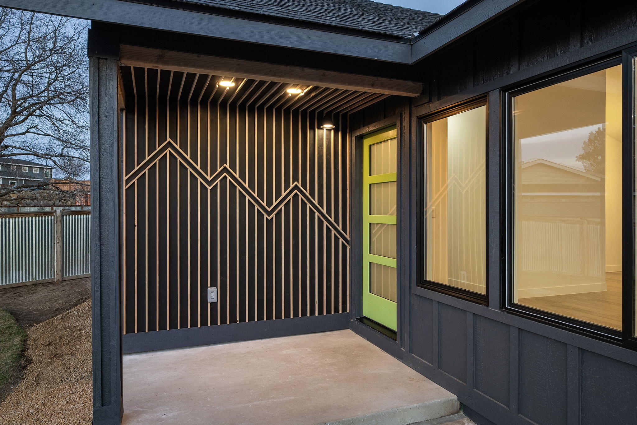 Close-up of modern building porch with black vertical wooden slats, a green door with three glass panels, and large windows, illuminated by two ceiling lights at dusk.