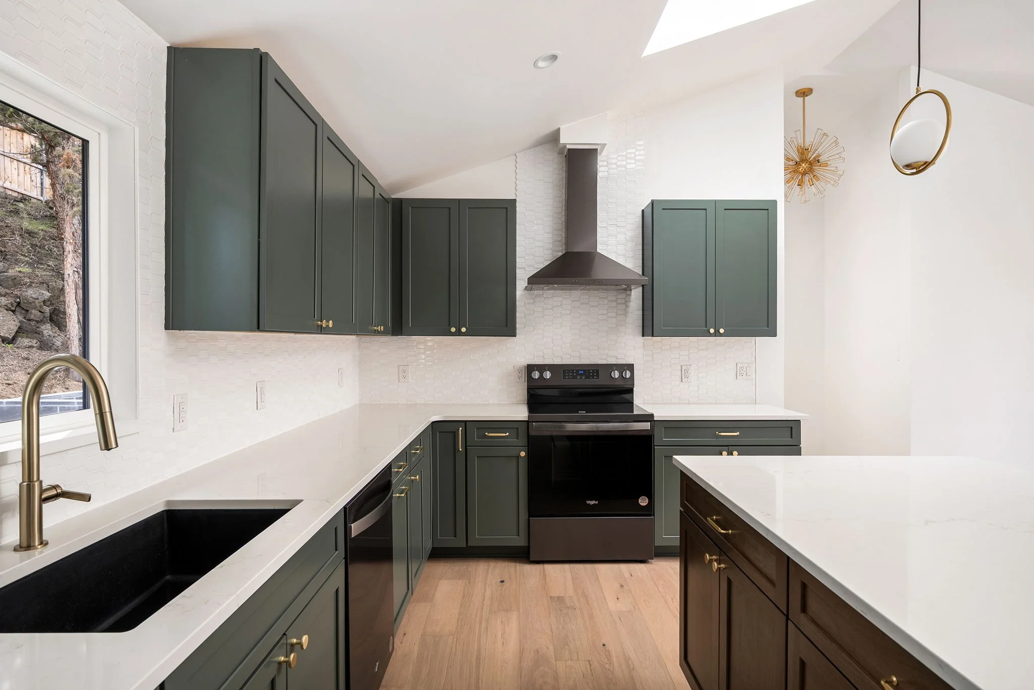 Modern kitchen with dark green cabinets, white countertops, black stove, stainless steel appliances, gold hardware, wooden flooring, and a window overlooking outdoor scenery.