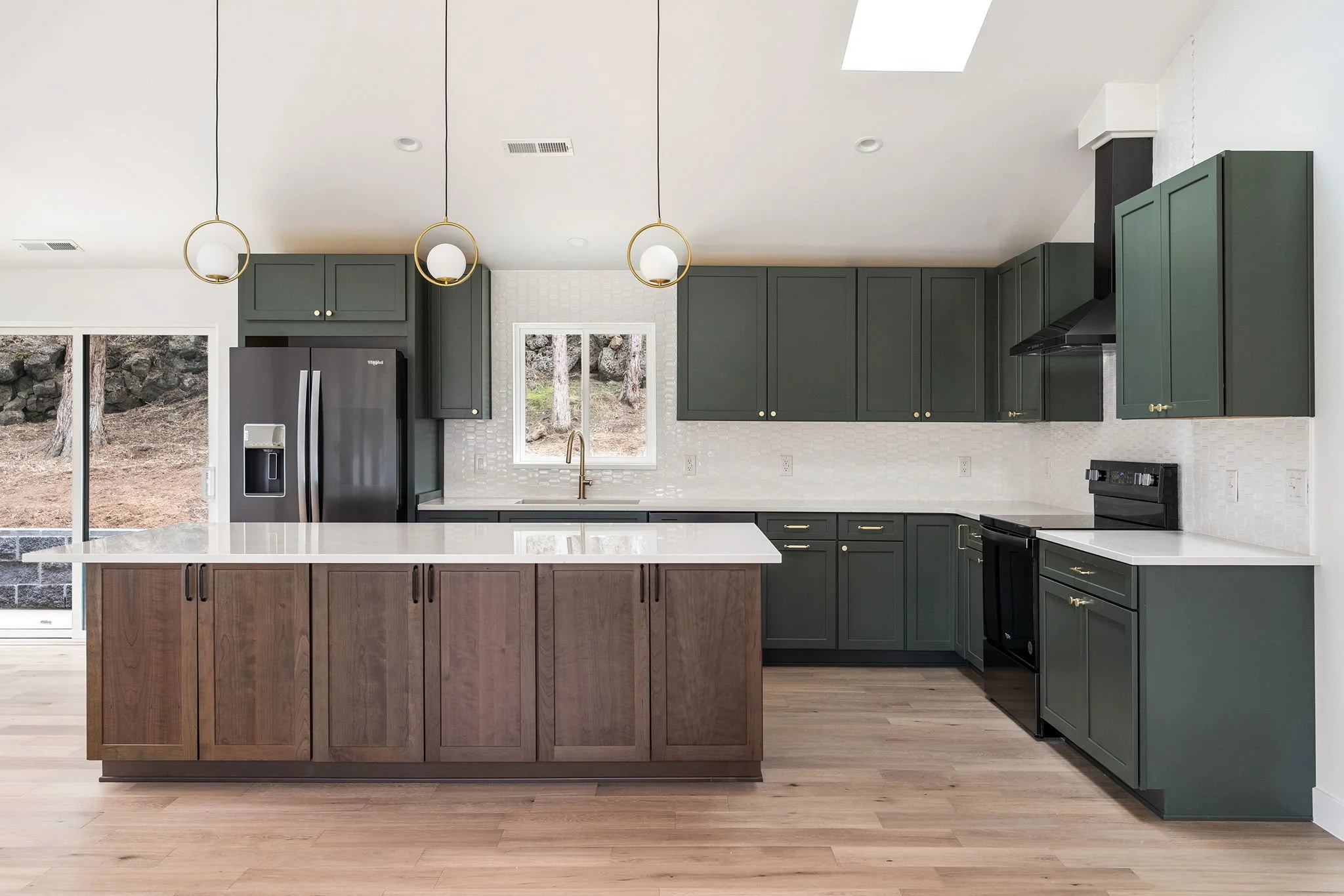 Modern kitchen with dark green cabinets, a white countertop, and a large wood-paneled island. Stainless steel appliances, a window above the sink, and a white tile backsplash. Hardwood floors and hanging pendant lights.