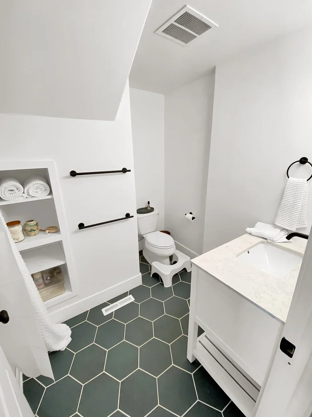 A modern bathroom with white walls, a white vanity with a marble countertop, a sink, black towel rings, a white toilet, and hexagonal dark gray tiles on the floor. There are black towel bars, an open shelf with towels and jars, and a small step stool