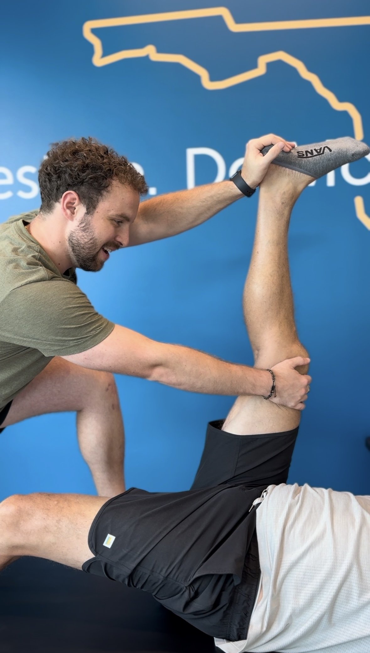 A Stretch Therapist assisting a patient with a leg stretch exercise in a gym with a blue backdrop featuring a yellow outline of the state of Florida.