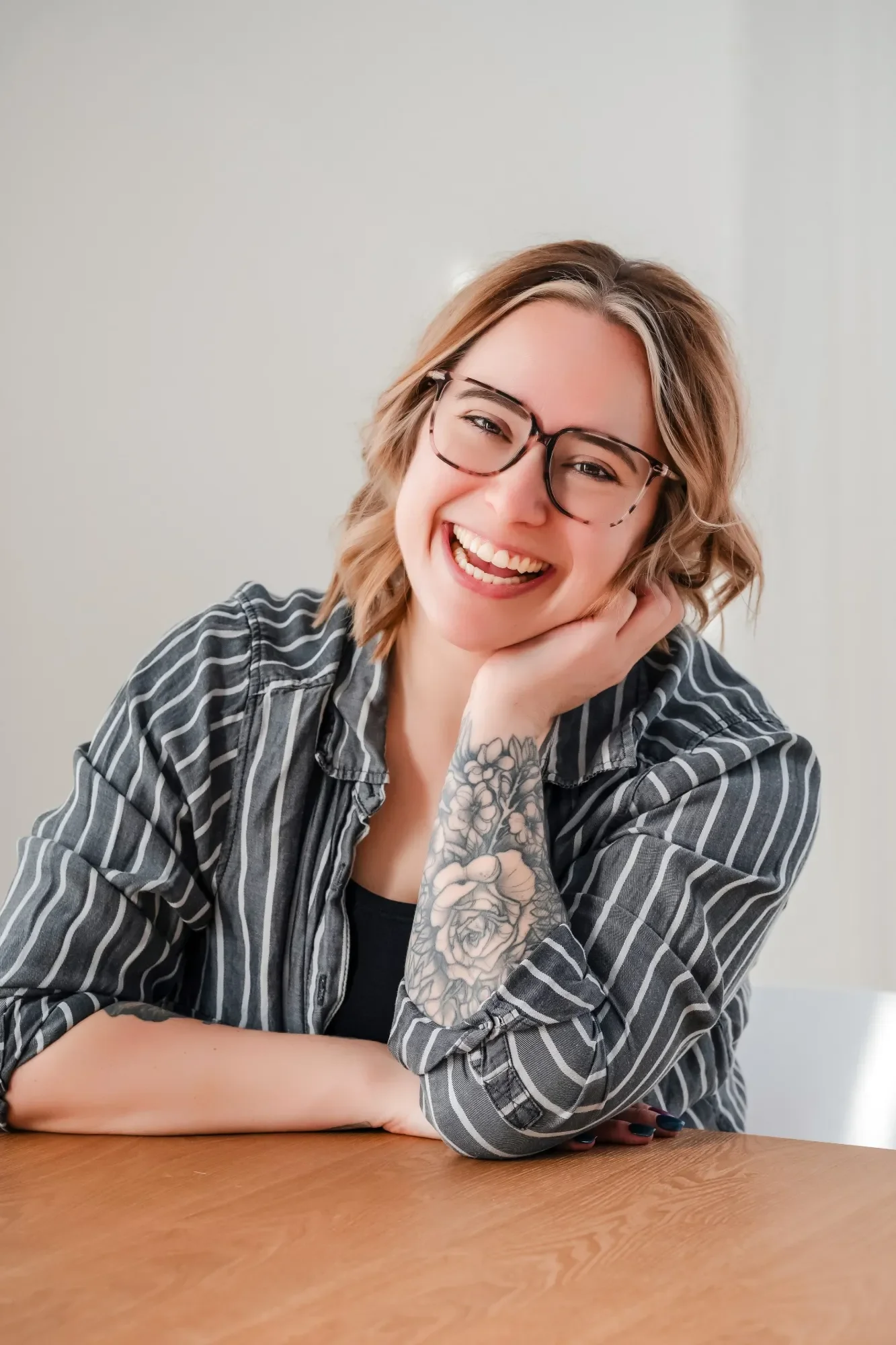 A smiling photo of photographer Samantha Turner with glasses, tattoos on her arm, and wavy blonde hair, sitting at a table with her hand resting on her cheek, wearing a striped shirt.