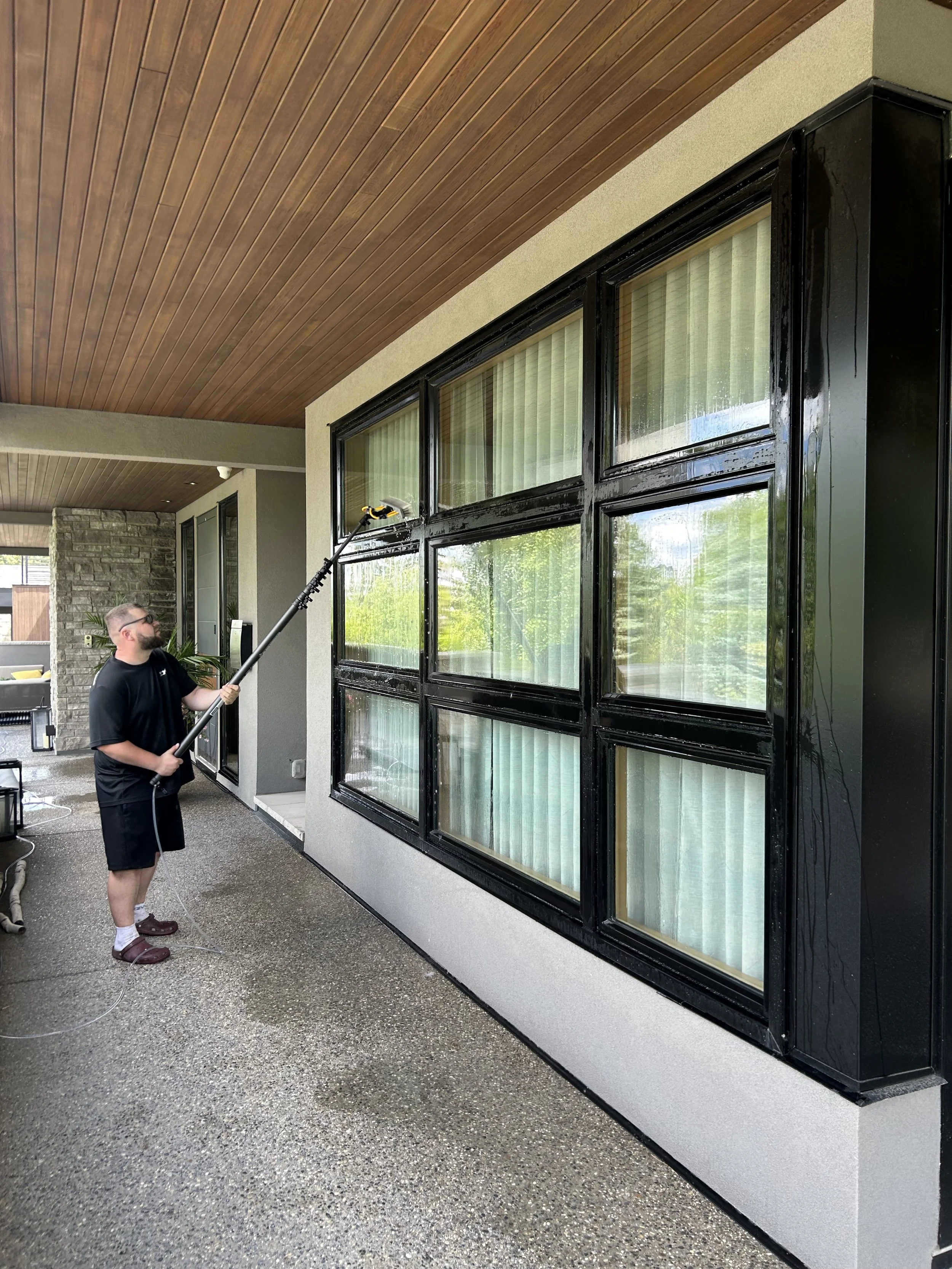 A man is cleaning the black-framed window of a house with a long-handled squeegee, standing on a wet concrete patio.