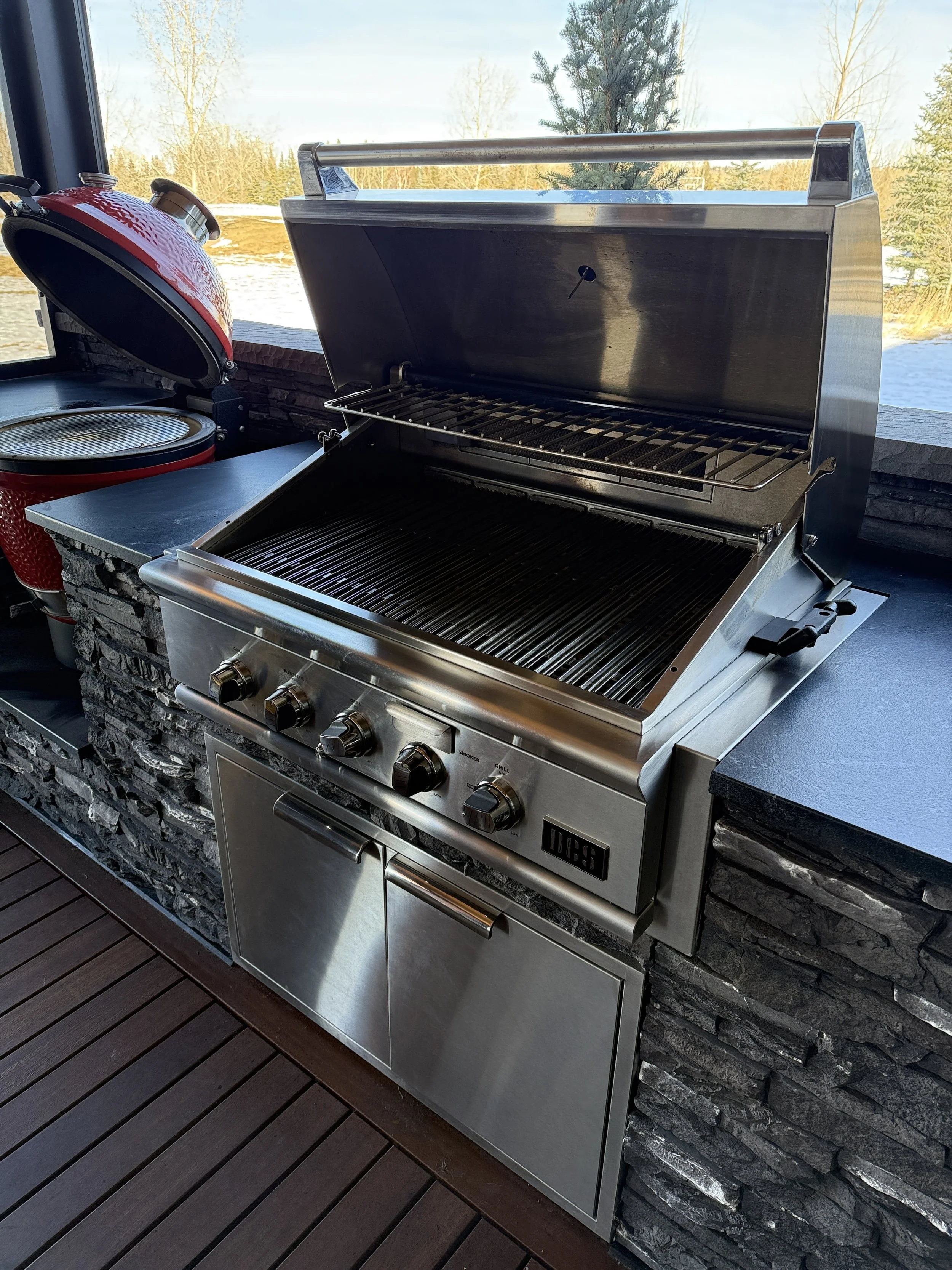 Outdoor kitchen with stainless steel gas grill, open lid, on a stone counter with a black countertop surface, overlooking snow-covered landscape with trees.