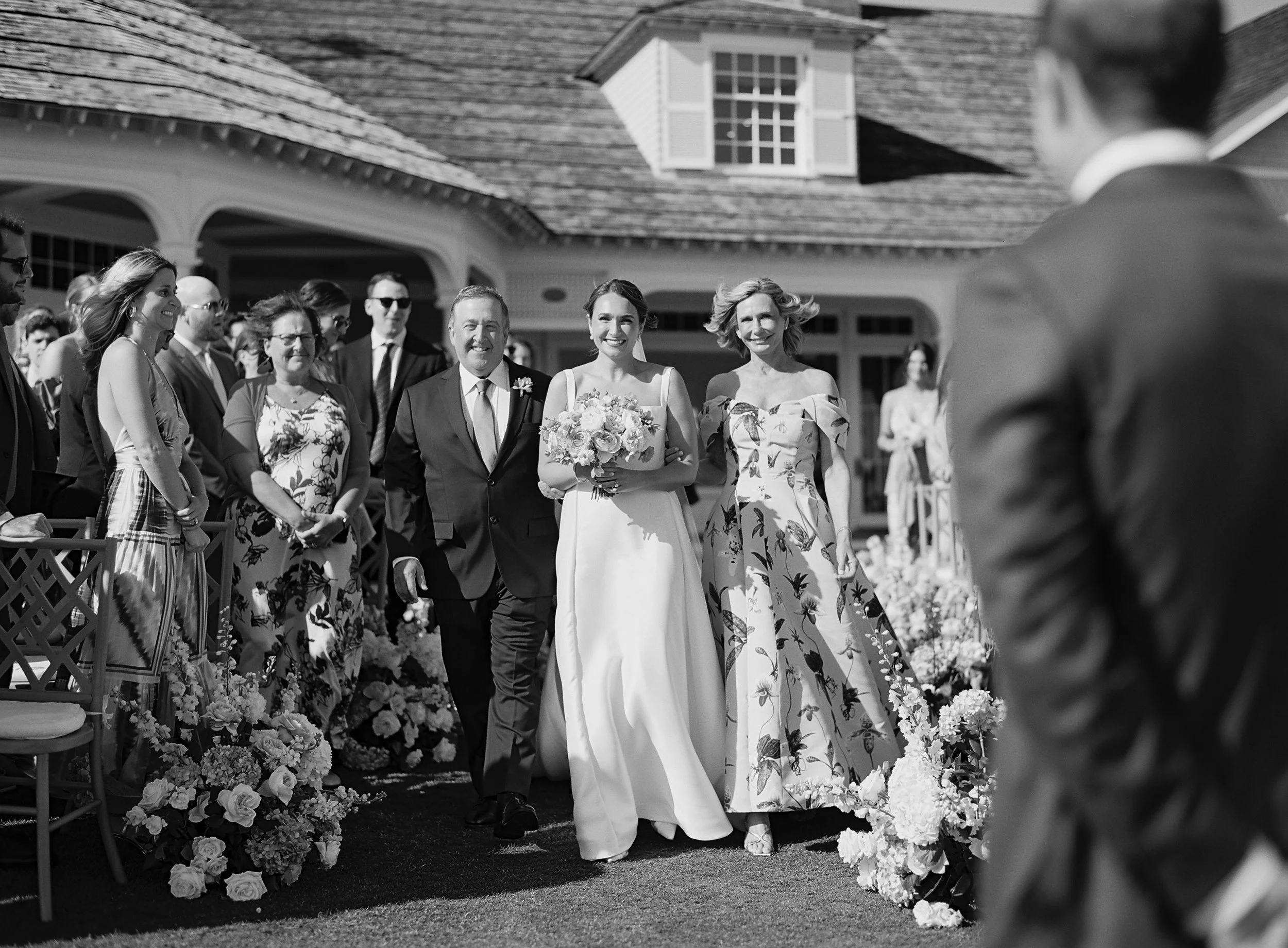 A bride walking down the aisle with her father, smiling and holding a bouquet, at an outdoor wedding with guests seated and standing on either side.