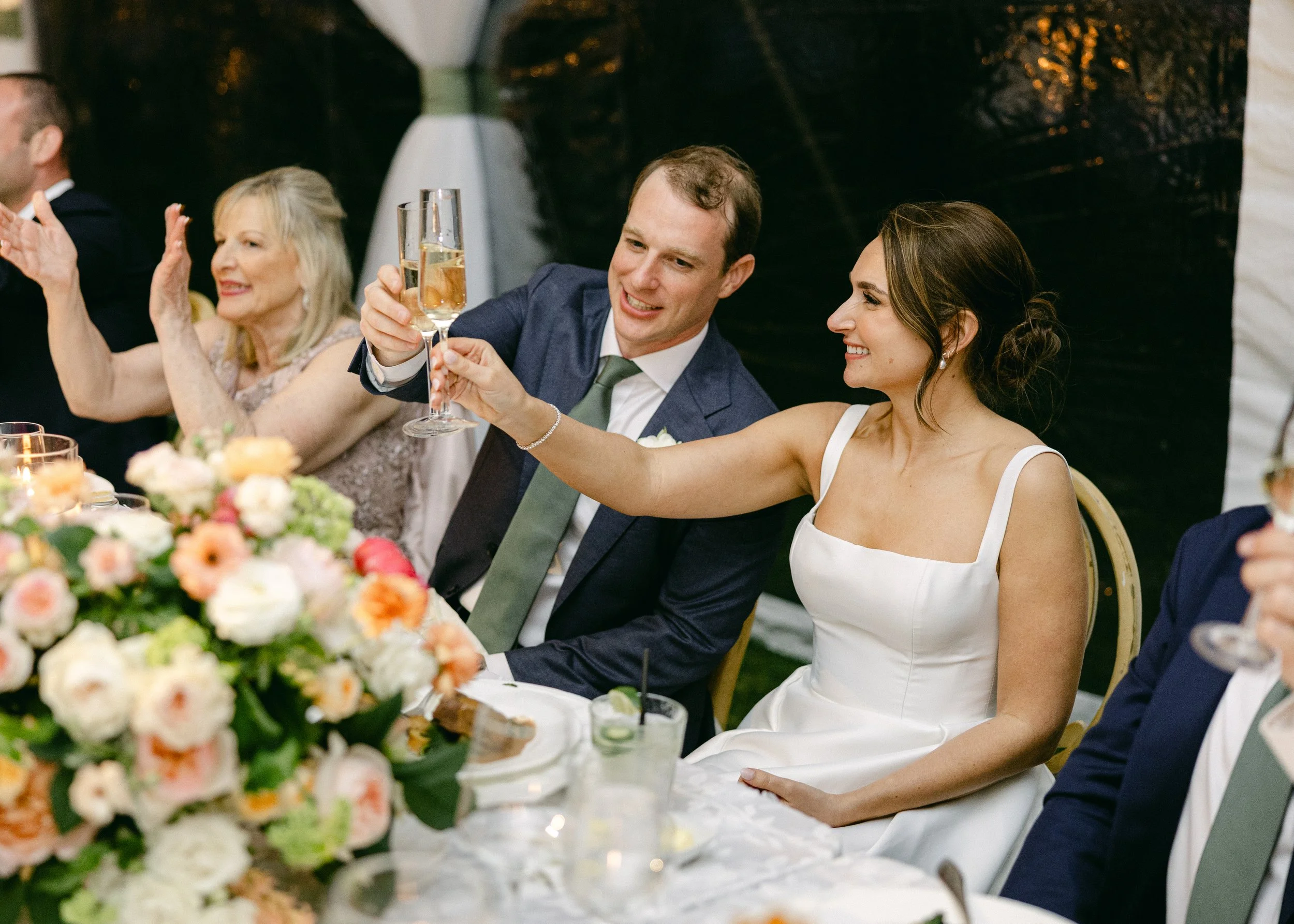 People raising glasses in celebration at a wedding reception, with a bride in a white dress and a groom in a blue suit.